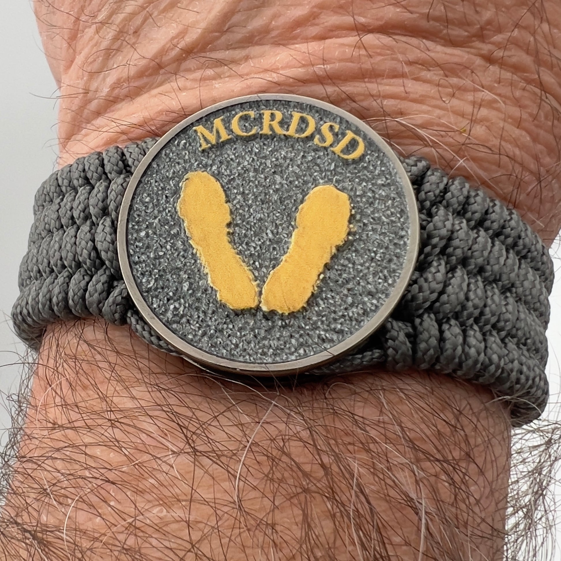 Thin, wide, flexible and comfortable braided United States Marine Corps Recruit Depot Yellow Footprints bracelet featuring a black nickel plated UV enamel painted medallion replica of an actual set of USMC footprints, a silver Cobra style tactical buckle and woven with Charcoal Gray Type-1 paracord.