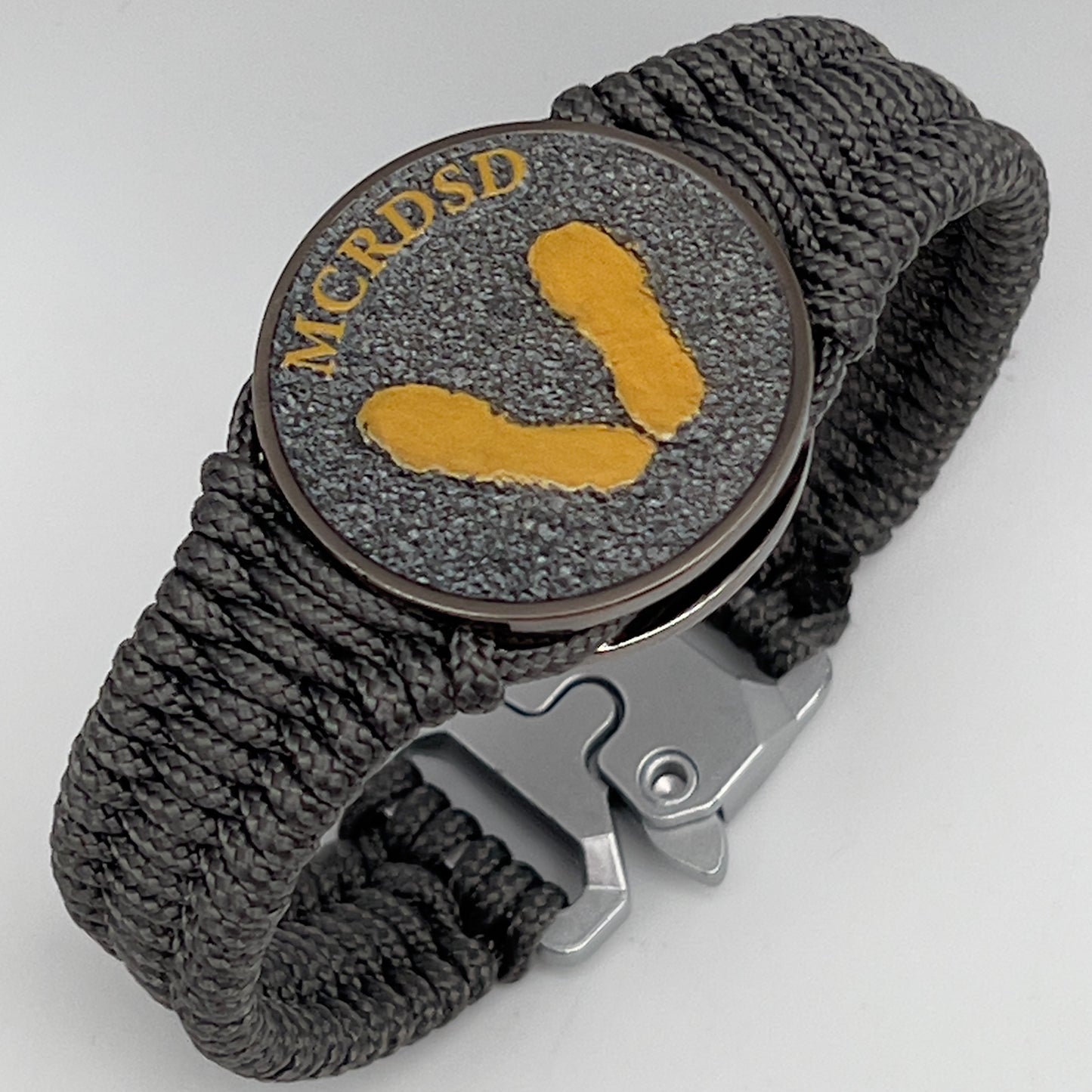 Thin, narrow, flexible and comfortable braided United States Marine Corps Recruit Depot Yellow Footprints bracelet featuring a black nickel plated UV enamel painted medallion replica of an actual set of USMC footprints, a silver Cobra style tactical buckle and woven with Charcoal Gray Type-1 paracord.