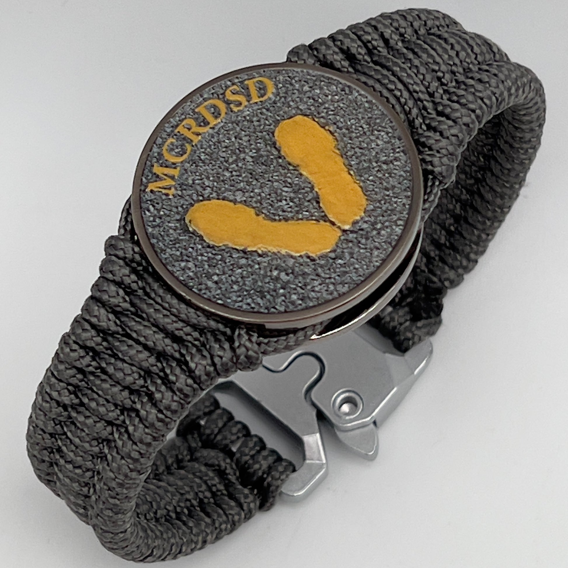Thin, narrow, flexible and comfortable braided United States Marine Corps Recruit Depot Yellow Footprints bracelet featuring a black nickel plated UV enamel painted medallion replica of an actual set of USMC footprints, a silver Cobra style tactical buckle and woven with Charcoal Gray Type-1 paracord.