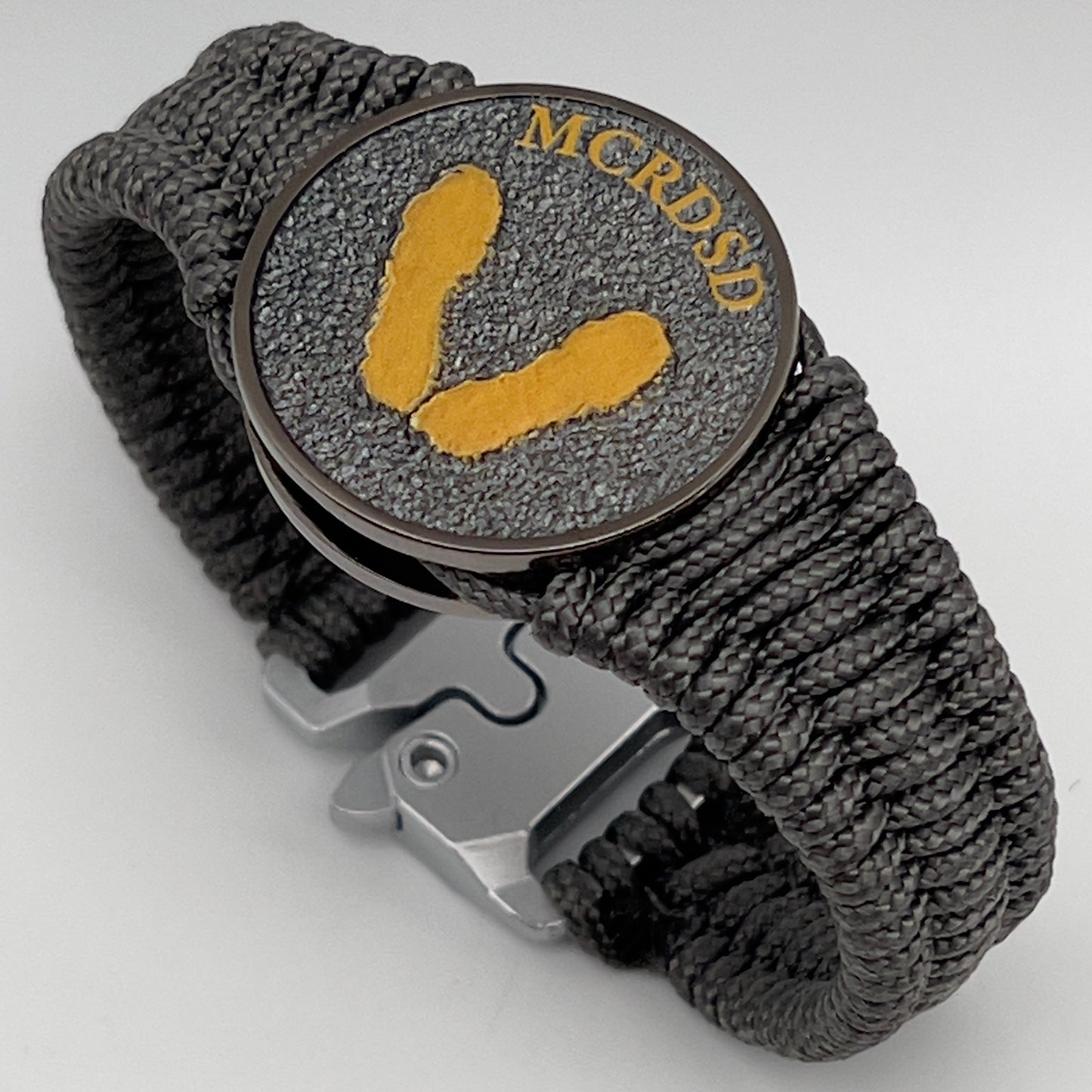 Thin, narrow, flexible and comfortable braided United States Marine Corps Recruit Depot Yellow Footprints bracelet featuring a black nickel plated UV enamel painted medallion replica of an actual set of USMC footprints, a silver Cobra style tactical buckle and woven with Charcoal Gray Type-1 paracord.