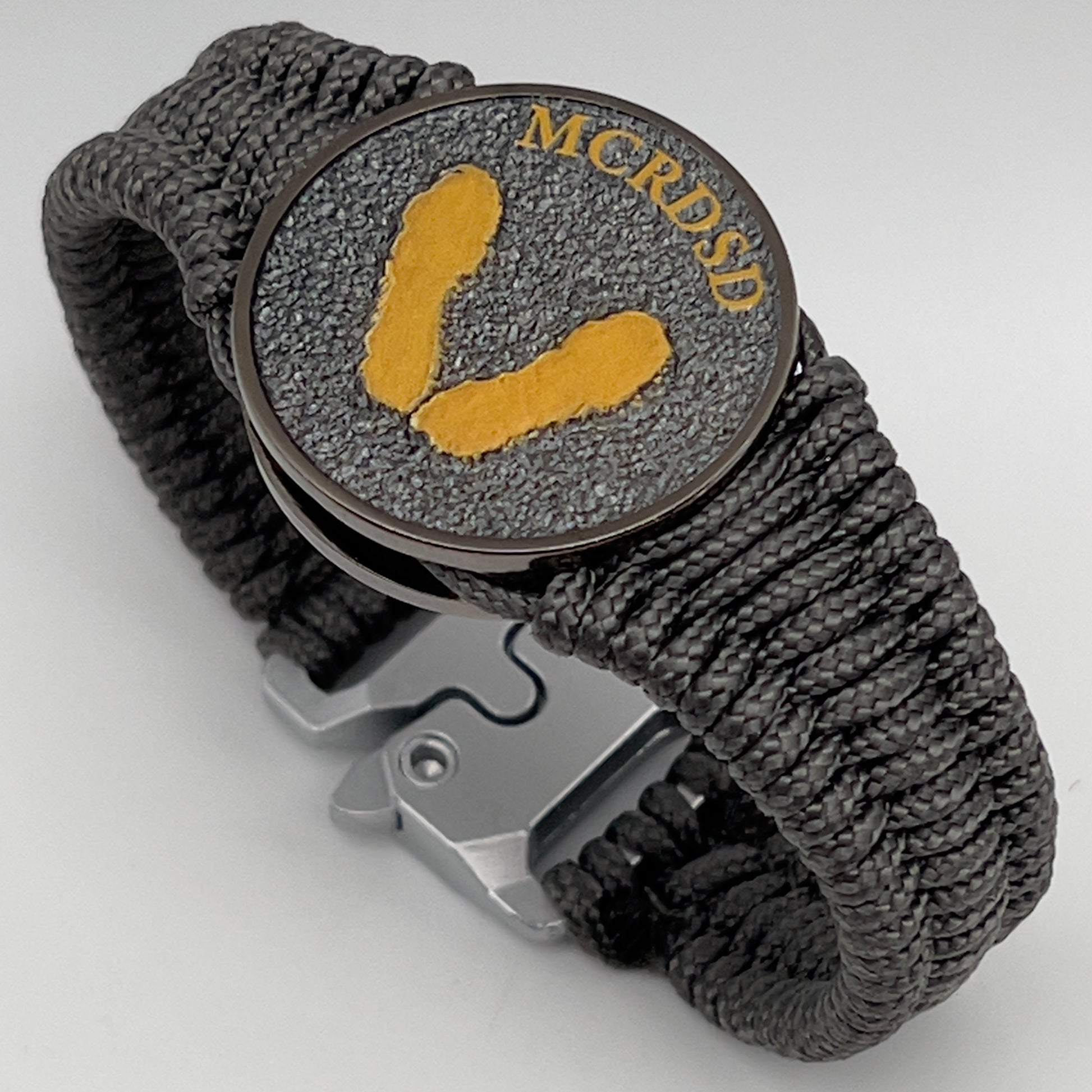 Thin, narrow, flexible and comfortable braided United States Marine Corps Recruit Depot Yellow Footprints bracelet featuring a black nickel plated UV enamel painted medallion replica of an actual set of USMC footprints, a silver Cobra style tactical buckle and woven with Charcoal Gray Type-1 paracord.