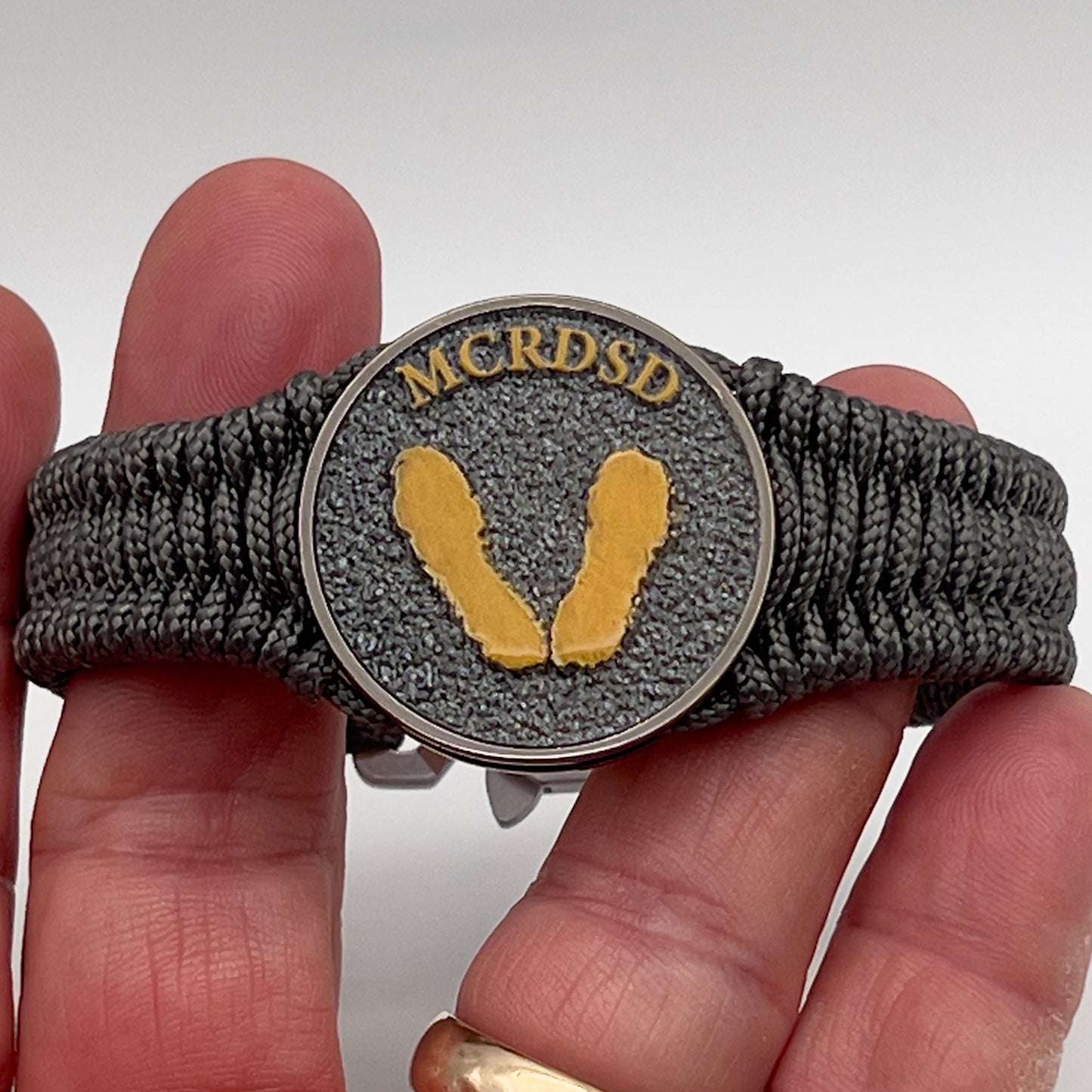 Thin, narrow, flexible and comfortable braided United States Marine Corps Recruit Depot Yellow Footprints bracelet featuring a black nickel plated UV enamel painted medallion replica of an actual set of USMC footprints, a silver Cobra style tactical buckle and woven with Charcoal Gray Type-1 paracord.