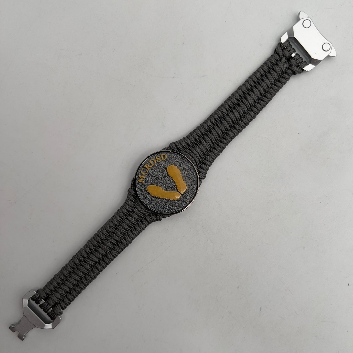 Thin, narrow, flexible and comfortable braided United States Marine Corps Recruit Depot Yellow Footprints bracelet featuring a black nickel plated UV enamel painted medallion replica of an actual set of USMC footprints, a silver Cobra style tactical buckle and woven with Charcoal Gray Type-1 paracord.