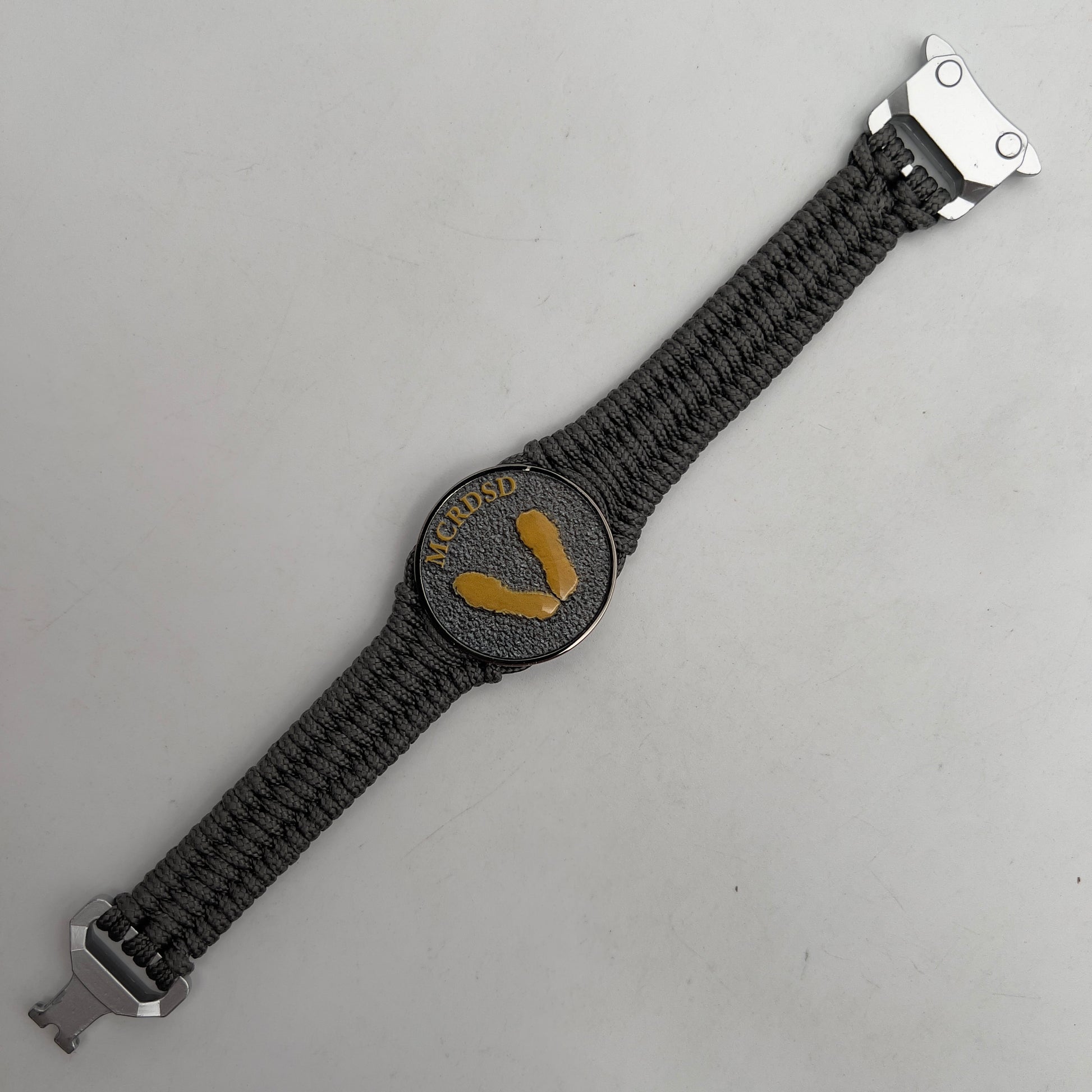 Thin, narrow, flexible and comfortable braided United States Marine Corps Recruit Depot Yellow Footprints bracelet featuring a black nickel plated UV enamel painted medallion replica of an actual set of USMC footprints, a silver Cobra style tactical buckle and woven with Charcoal Gray Type-1 paracord.