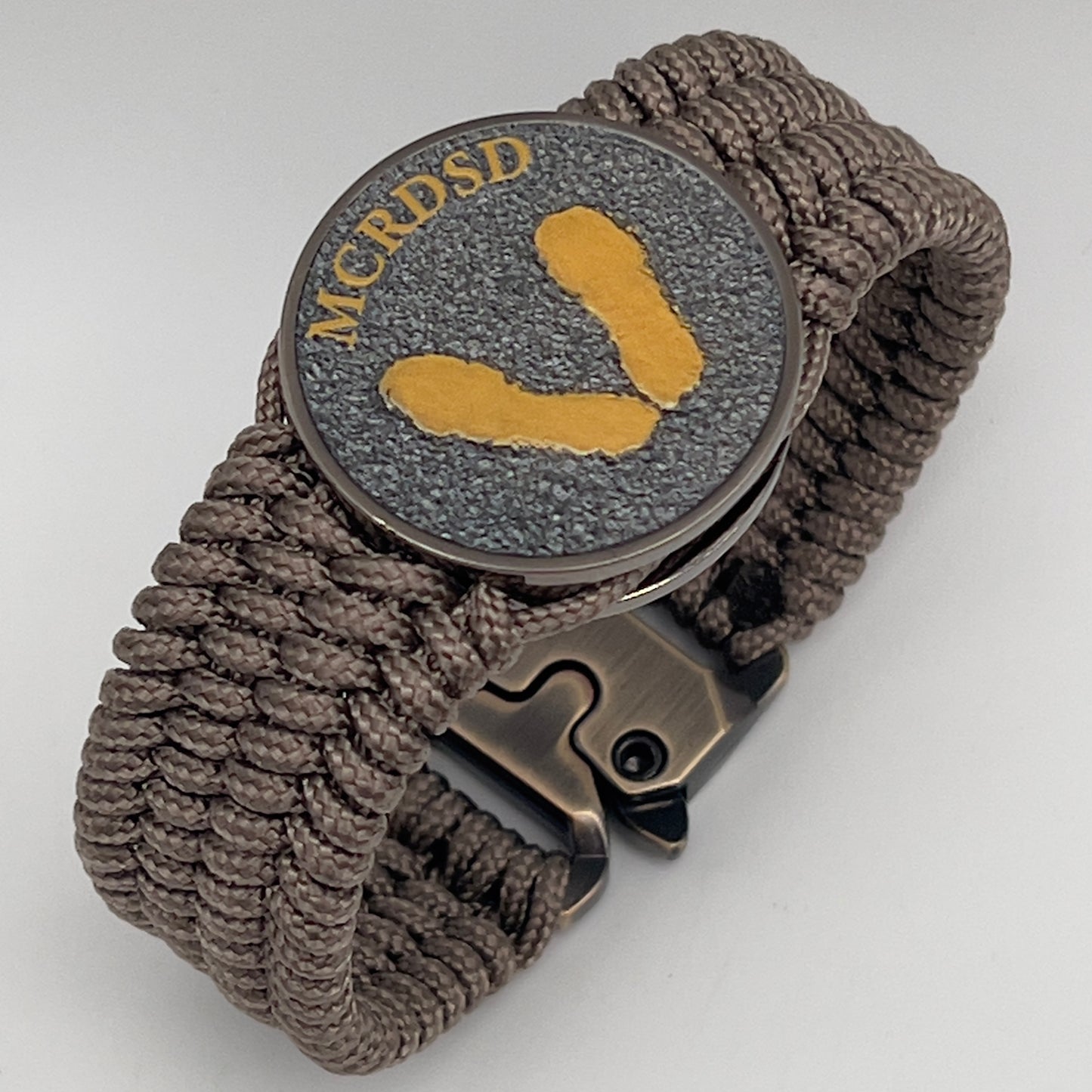 Thin, wide, flexible and comfortable braided United States Marine Corps Recruit Depot Yellow Footprints bracelet featuring a black nickel plated UV enamel painted medallion replica of an actual set of USMC recruit depot footprints, a gold Cobra style tactical buckle and woven with tan Type-1 paracord.