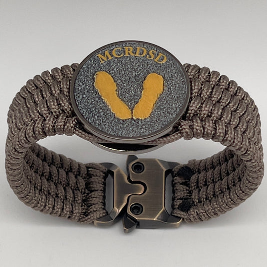 Thin, wide, flexible and comfortable braided United States Marine Corps Recruit Depot Yellow Footprints bracelet featuring a black nickel plated UV enamel painted medallion replica of an actual set of USMC recruit depot footprints, a gold Cobra style tactical buckle and woven with tan Type-1 paracord.