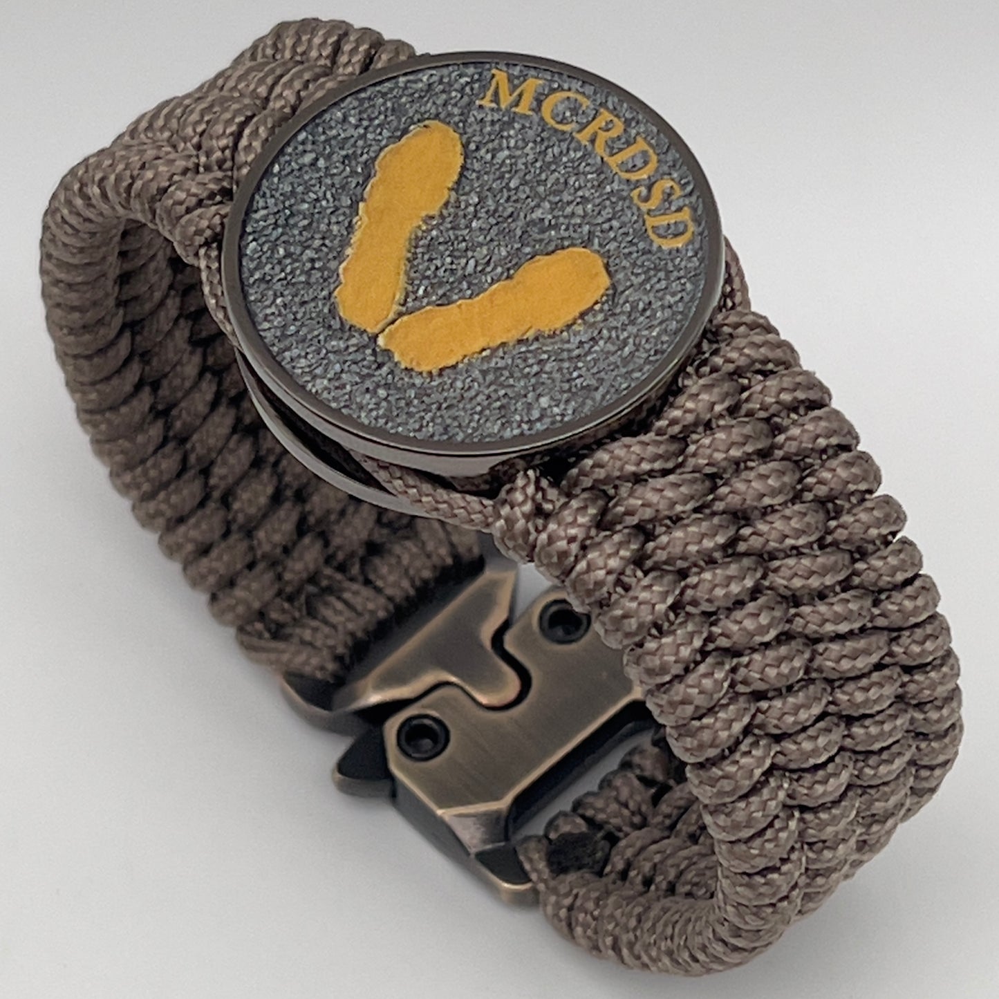 Thin, wide, flexible and comfortable braided United States Marine Corps Recruit Depot Yellow Footprints bracelet featuring a black nickel plated UV enamel painted medallion replica of an actual set of USMC recruit depot footprints, a gold Cobra style tactical buckle and woven with tan Type-1 paracord.