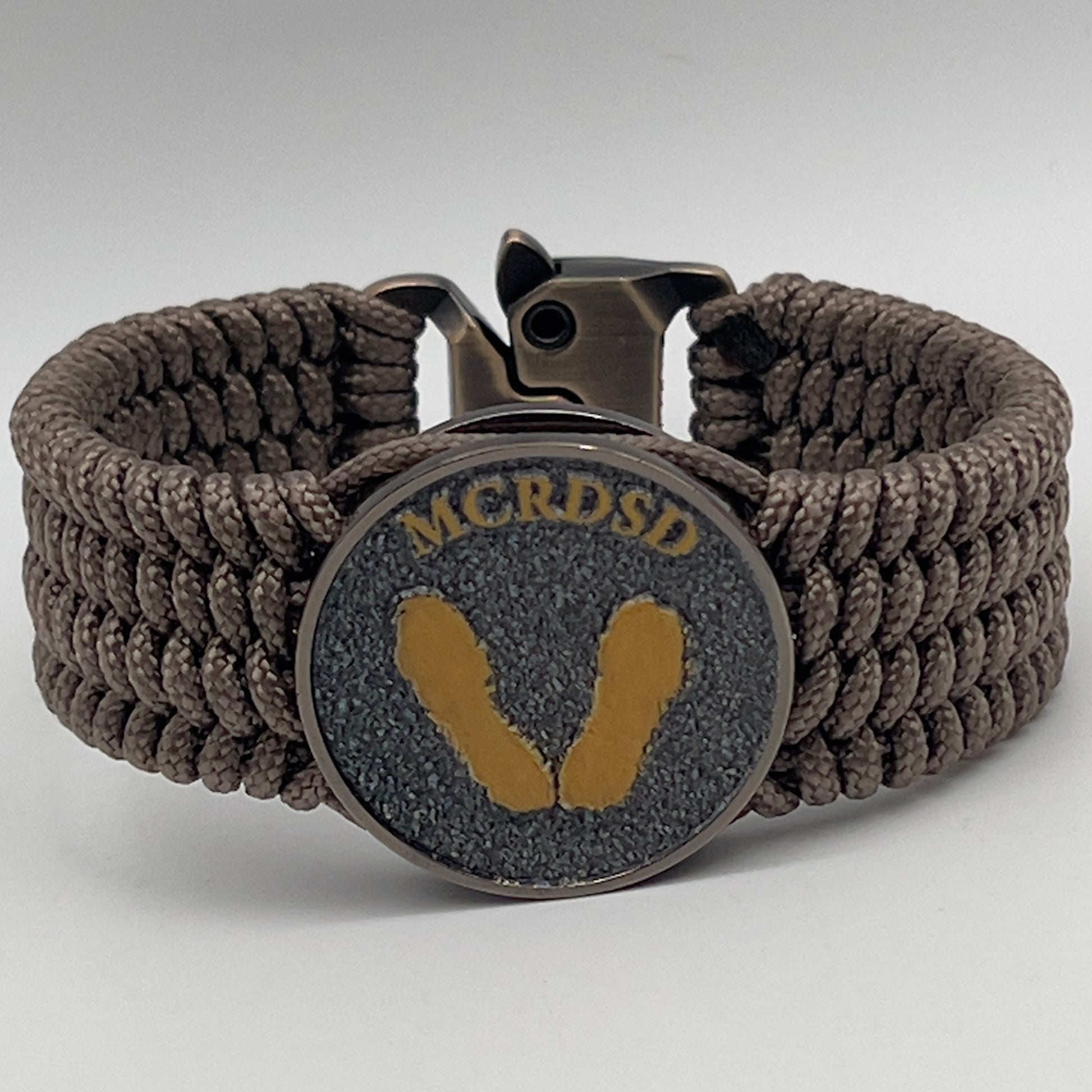 Thin, wide, flexible and comfortable braided United States Marine Corps Recruit Depot Yellow Footprints bracelet featuring a black nickel plated UV enamel painted medallion replica of an actual set of USMC recruit depot footprints, a gold Cobra style tactical buckle and woven with tan Type-1 paracord.