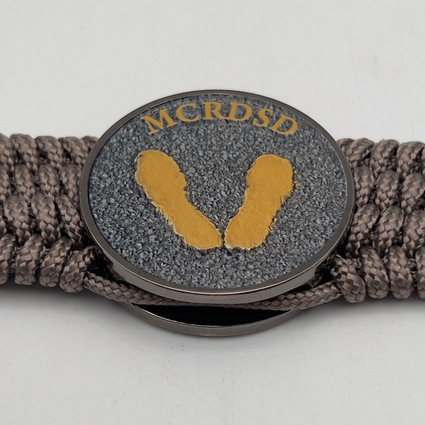 Thin, wide, flexible and comfortable braided United States Marine Corps Recruit Depot Yellow Footprints bracelet featuring a black nickel plated UV enamel painted medallion replica of an actual set of USMC recruit depot footprints, a gold Cobra style tactical buckle and woven with tan Type-1 paracord.