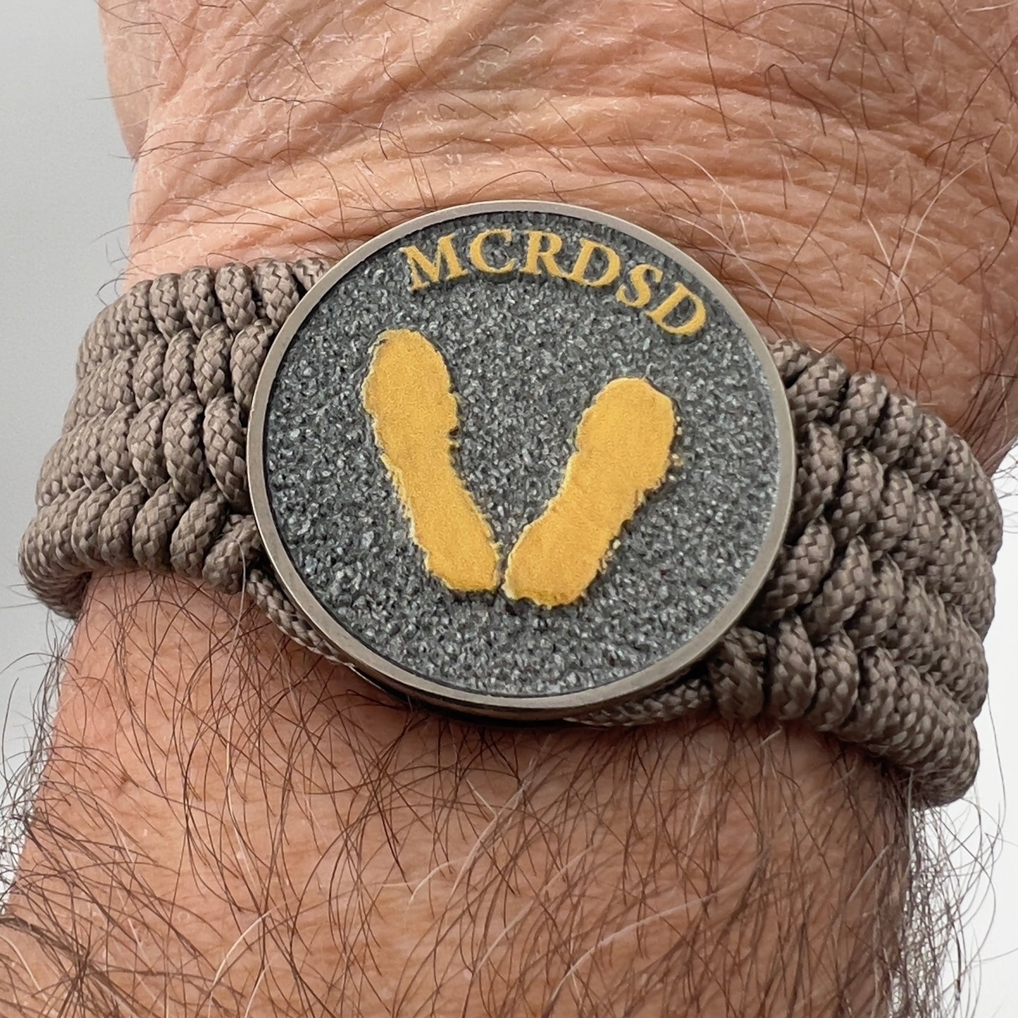 Thin, wide, flexible and comfortable braided United States Marine Corps Recruit Depot Yellow Footprints bracelet featuring a black nickel plated UV enamel painted medallion replica of an actual set of USMC recruit depot footprints, a gold Cobra style tactical buckle and woven with tan Type-1 paracord.