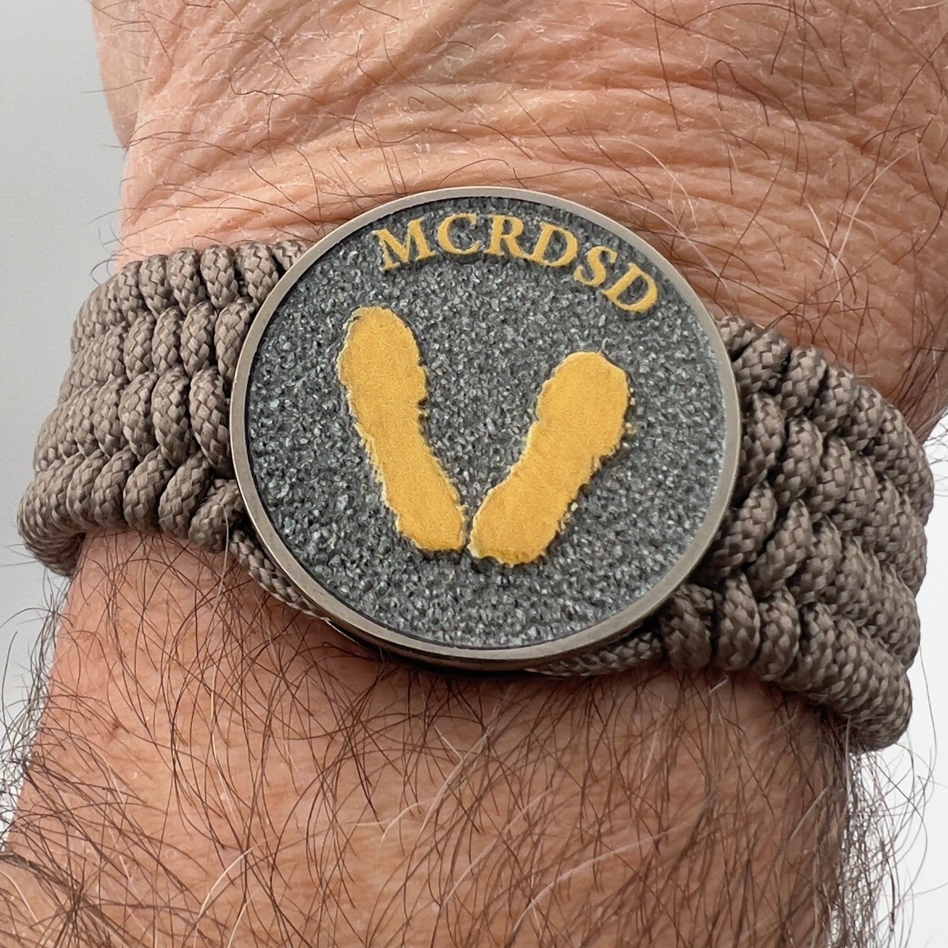Thin, wide, flexible and comfortable braided United States Marine Corps Recruit Depot Yellow Footprints bracelet featuring a black nickel plated UV enamel painted medallion replica of an actual set of USMC recruit depot footprints, a gold Cobra style tactical buckle and woven with tan Type-1 paracord.
