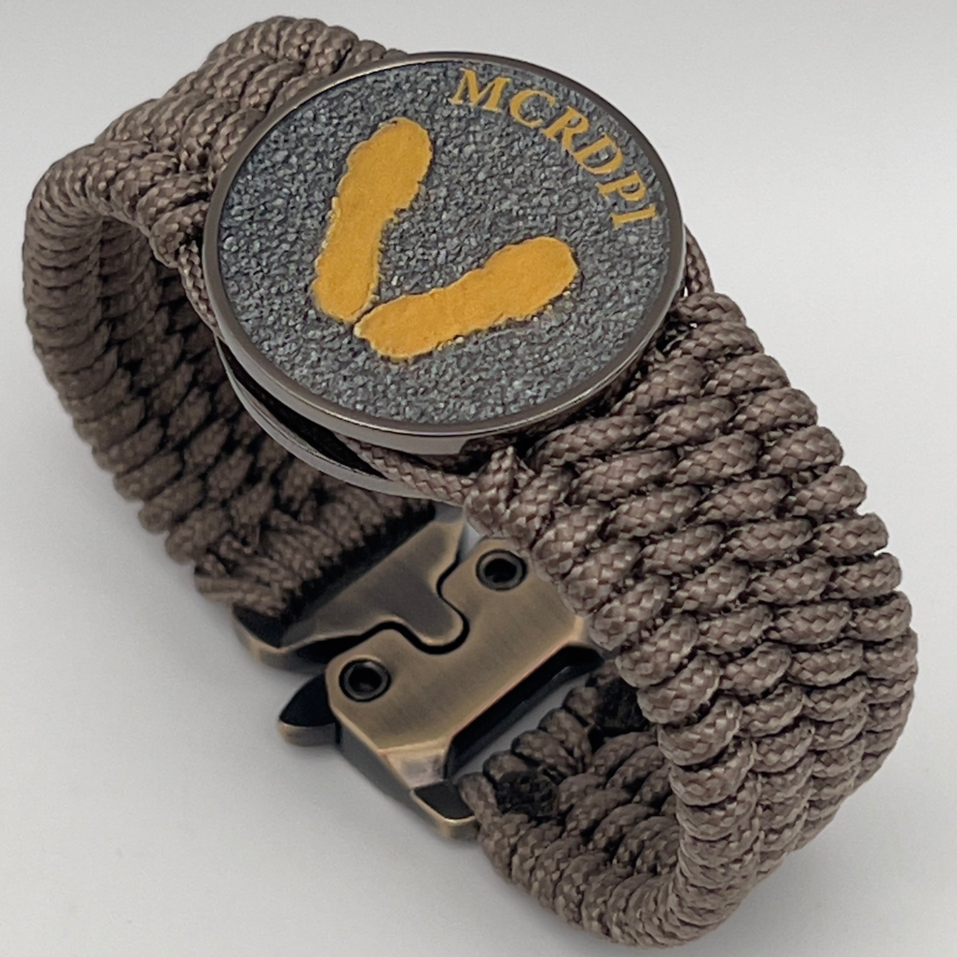 Thin, wide, flexible and comfortable braided United States Marine Corps Recruit Depot Yellow Footprints bracelet featuring a black nickel plated UV enamel painted medallion replica of an actual set of USMC recruit depot footprints, a gold Cobra style tactical buckle and woven with tan Type-1 paracord.
