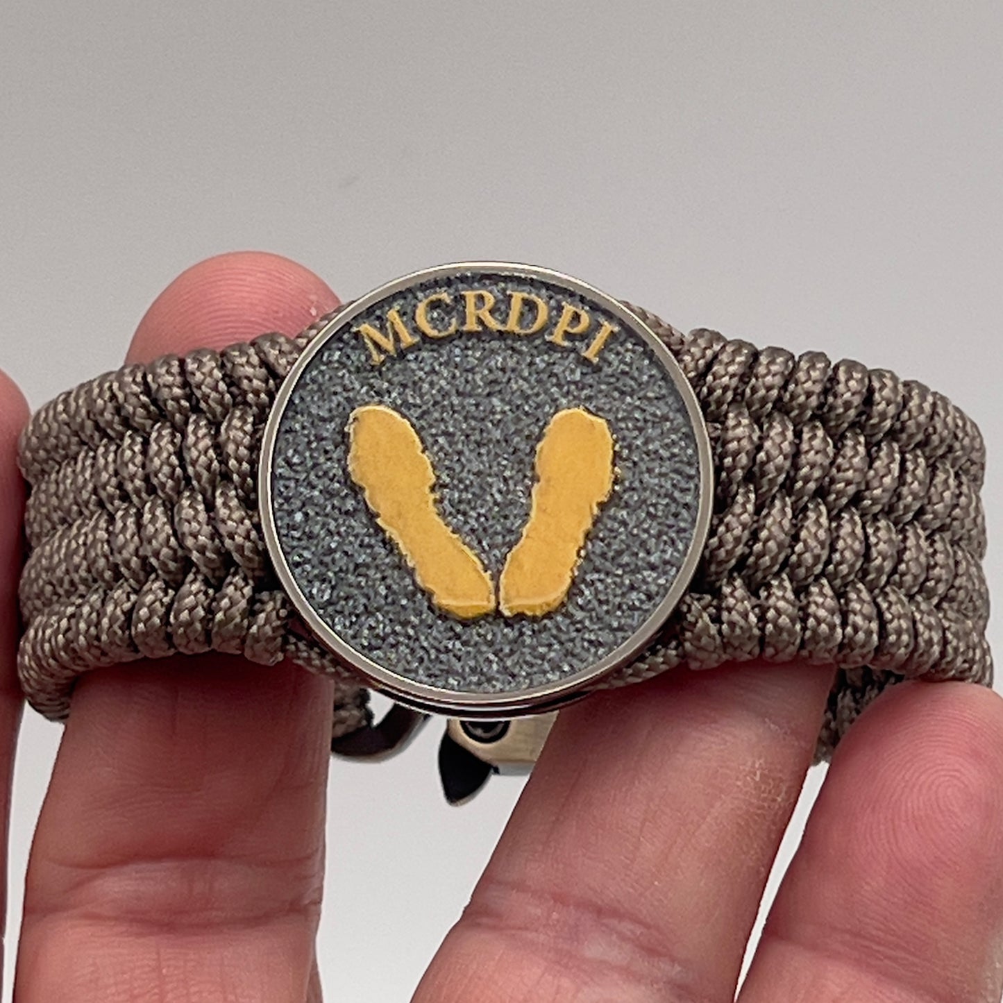 Thin, wide, flexible and comfortable braided United States Marine Corps Recruit Depot Yellow Footprints bracelet featuring a black nickel plated UV enamel painted medallion replica of an actual set of USMC recruit depot footprints, a gold Cobra style tactical buckle and woven with tan Type-1 paracord.