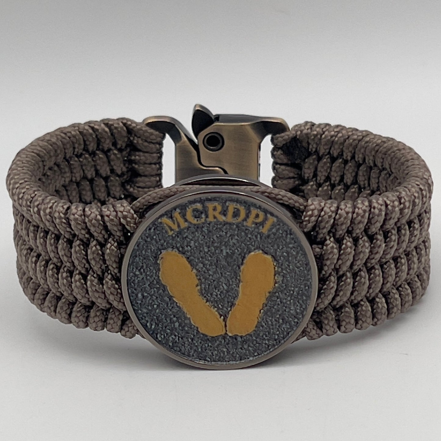 Thin, wide, flexible and comfortable braided United States Marine Corps Recruit Depot Yellow Footprints bracelet featuring a black nickel plated UV enamel painted medallion replica of an actual set of USMC recruit depot footprints, a gold Cobra style tactical buckle and woven with tan Type-1 paracord.