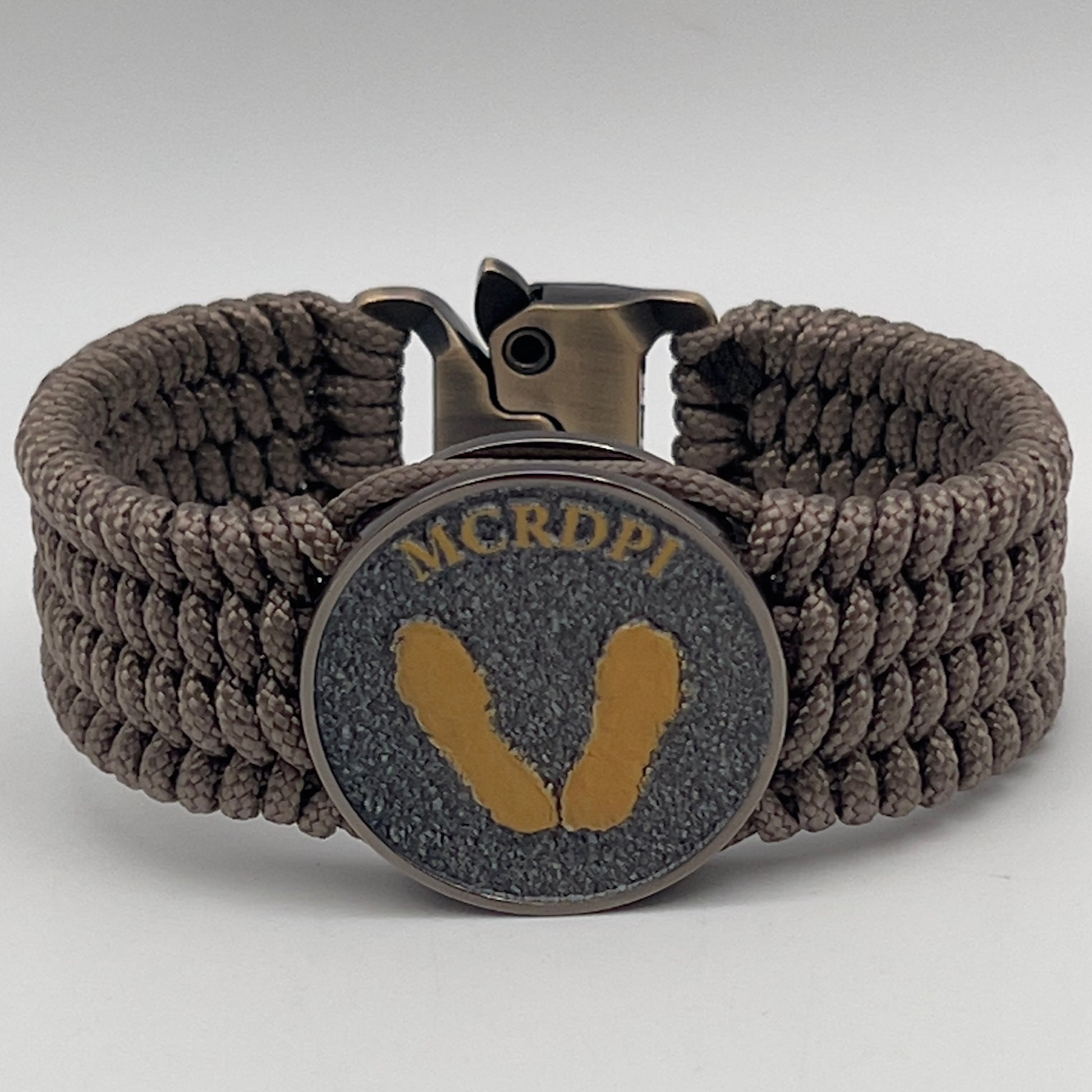 Thin, wide, flexible and comfortable braided United States Marine Corps Recruit Depot Yellow Footprints bracelet featuring a black nickel plated UV enamel painted medallion replica of an actual set of USMC recruit depot footprints, a gold Cobra style tactical buckle and woven with tan Type-1 paracord.