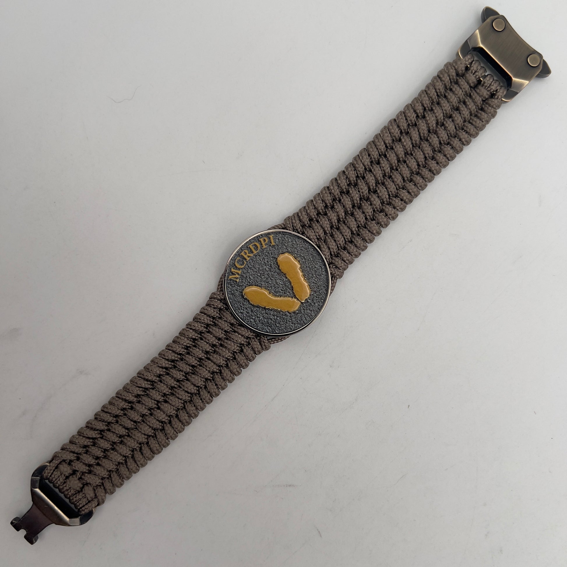 Thin, wide, flexible and comfortable braided United States Marine Corps Recruit Depot Yellow Footprints bracelet featuring a black nickel plated UV enamel painted medallion replica of an actual set of USMC recruit depot footprints, a gold Cobra style tactical buckle and woven with tan Type-1 paracord.
