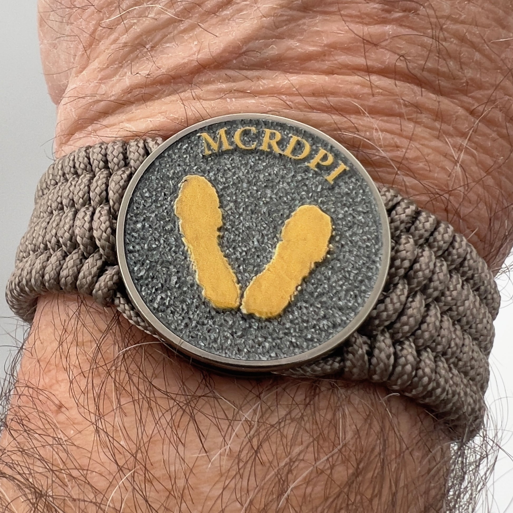 Thin, wide, flexible and comfortable braided United States Marine Corps Recruit Depot Yellow Footprints bracelet featuring a black nickel plated UV enamel painted medallion replica of an actual set of USMC recruit depot footprints, a gold Cobra style tactical buckle and woven with tan Type-1 paracord.