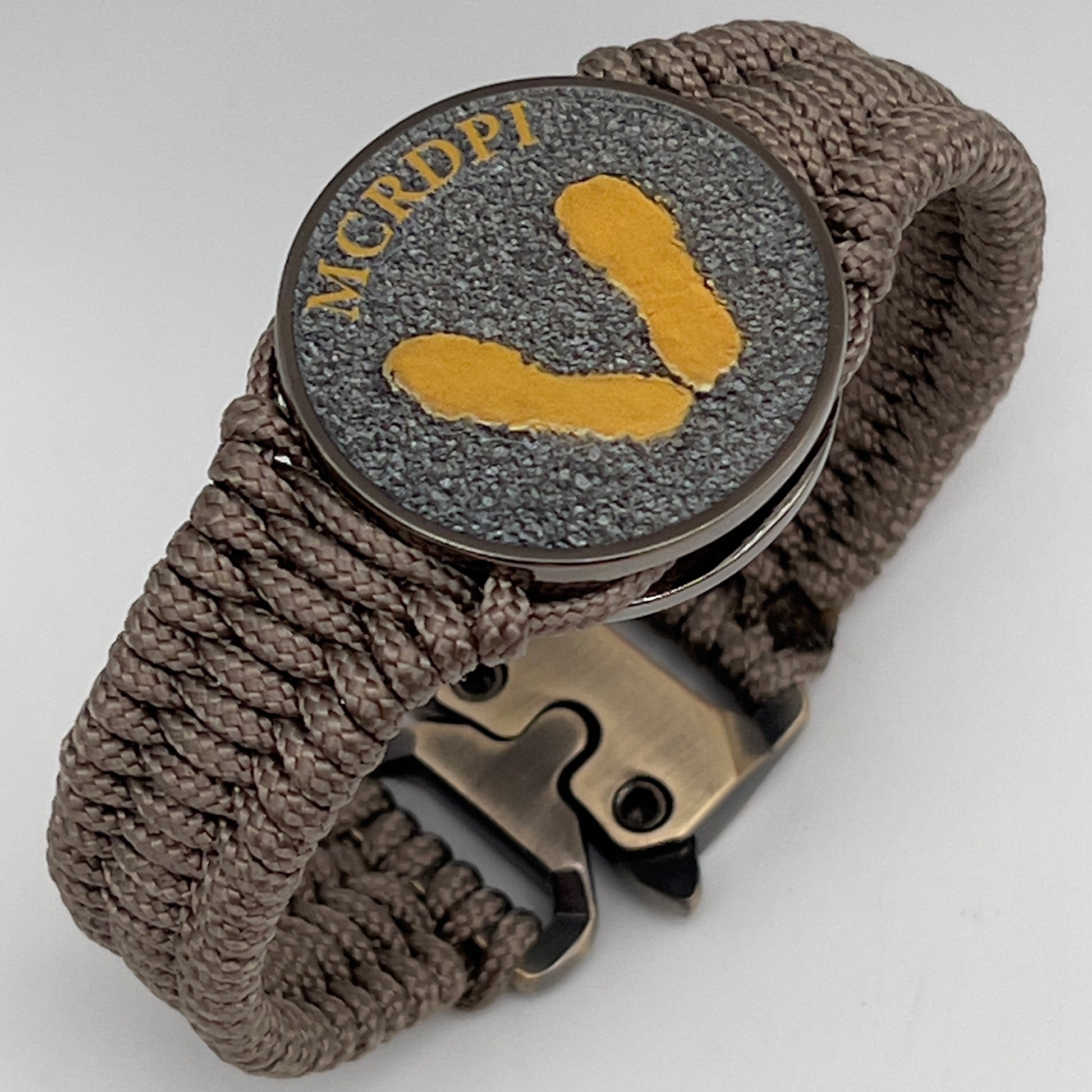 Thin, narrow, flexible and comfortable braided United States Marine Corps Recruit Depot Yellow Footprints bracelet featuring a black nickel plated UV enamel painted medallion replica of an actual set of USMC Yellow Footprints footprints, a gold Cobra style tactical buckle and woven with tan Type-1 paracord.