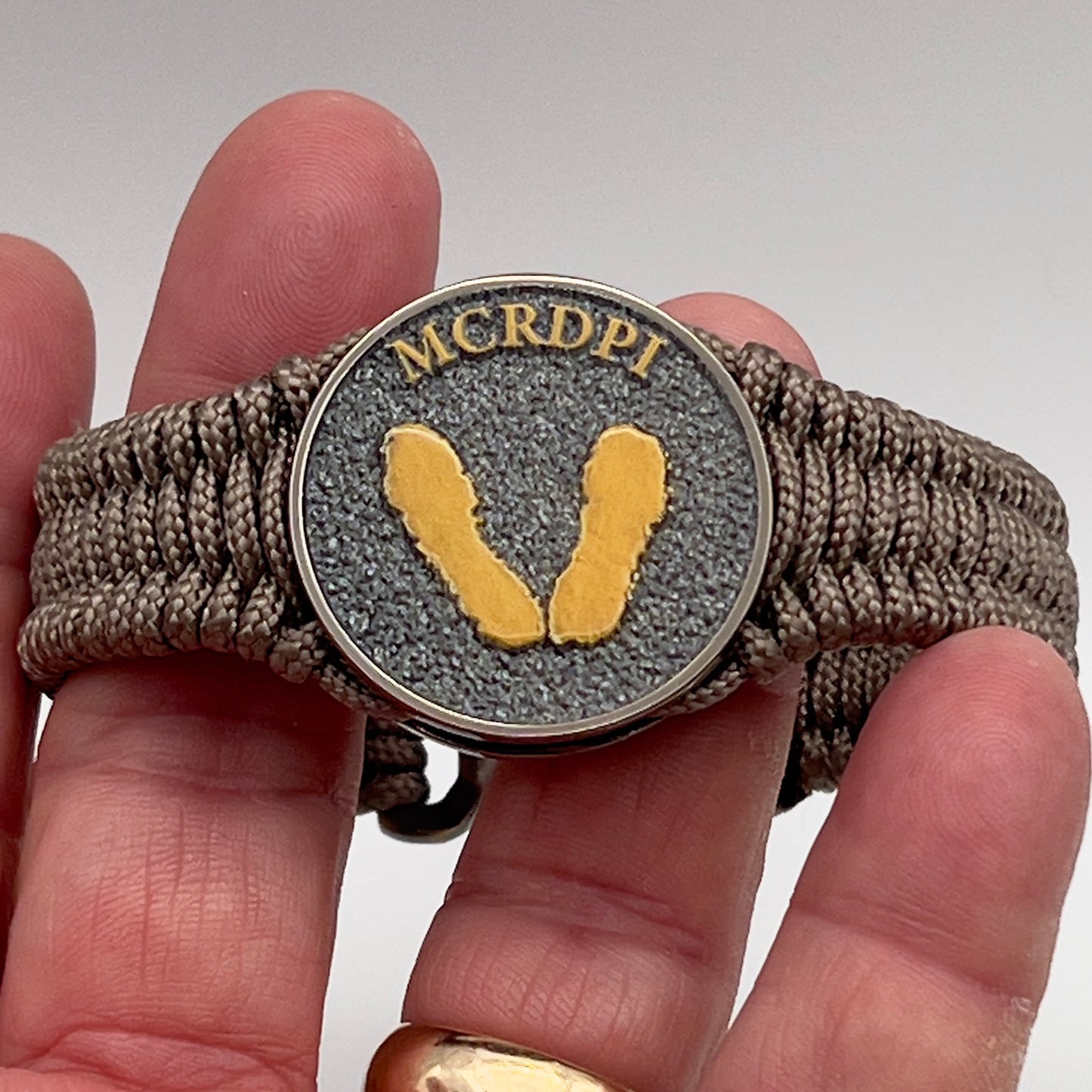 Thin, narrow, flexible and comfortable braided United States Marine Corps Recruit Depot Yellow Footprints bracelet featuring a black nickel plated UV enamel painted medallion replica of an actual set of USMC Yellow Footprints footprints, a gold Cobra style tactical buckle and woven with tan Type-1 paracord.