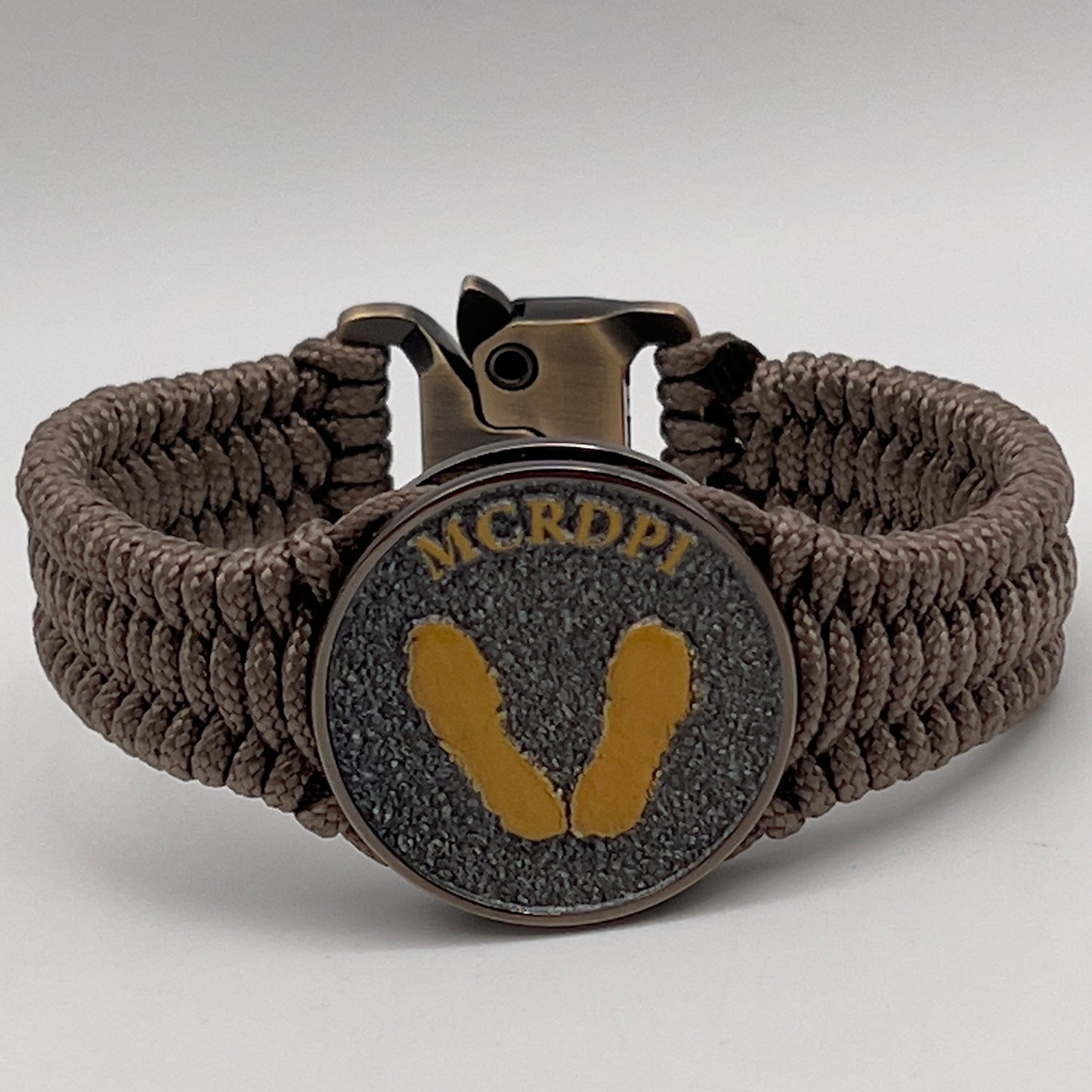 Thin, narrow, flexible and comfortable braided United States Marine Corps Recruit Depot Yellow Footprints bracelet featuring a black nickel plated UV enamel painted medallion replica of an actual set of USMC Yellow Footprints footprints, a gold Cobra style tactical buckle and woven with tan Type-1 paracord.
