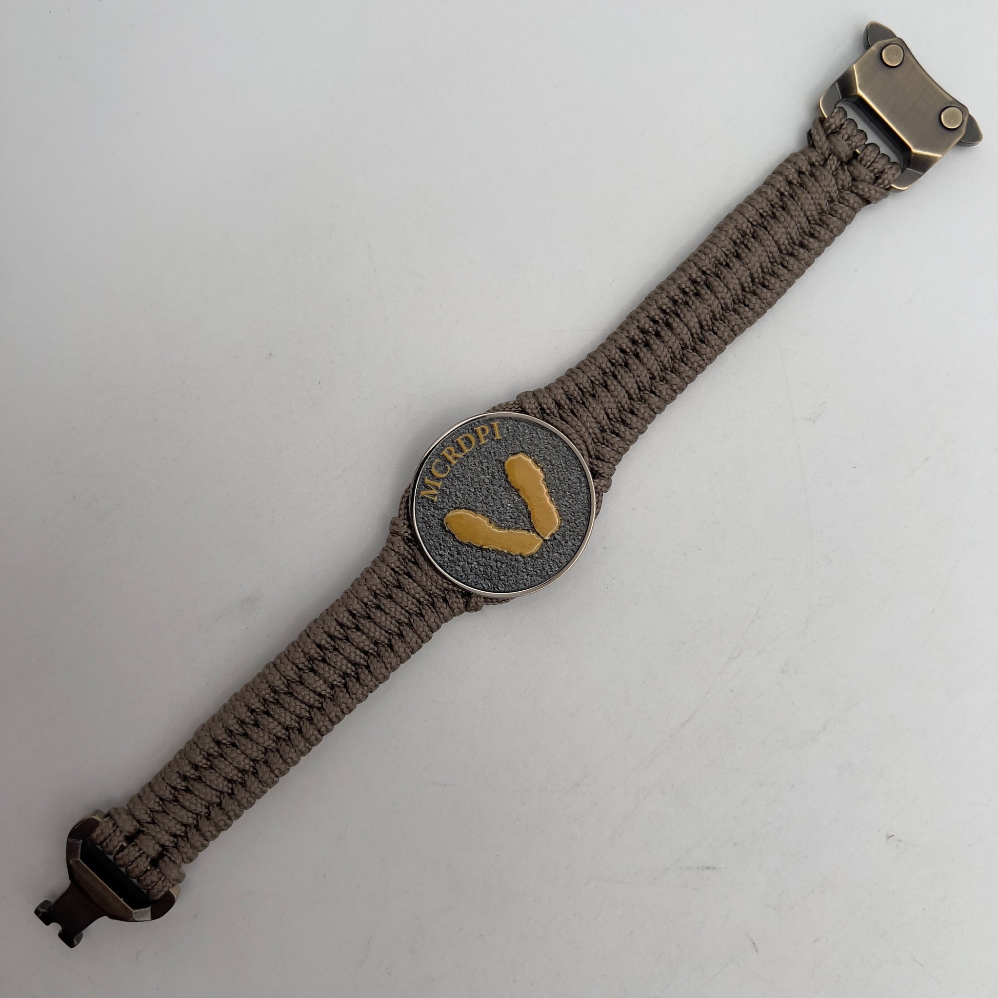 Thin, narrow, flexible and comfortable braided United States Marine Corps Recruit Depot Yellow Footprints bracelet featuring a black nickel plated UV enamel painted medallion replica of an actual set of USMC Yellow Footprints footprints, a gold Cobra style tactical buckle and woven with tan Type-1 paracord.