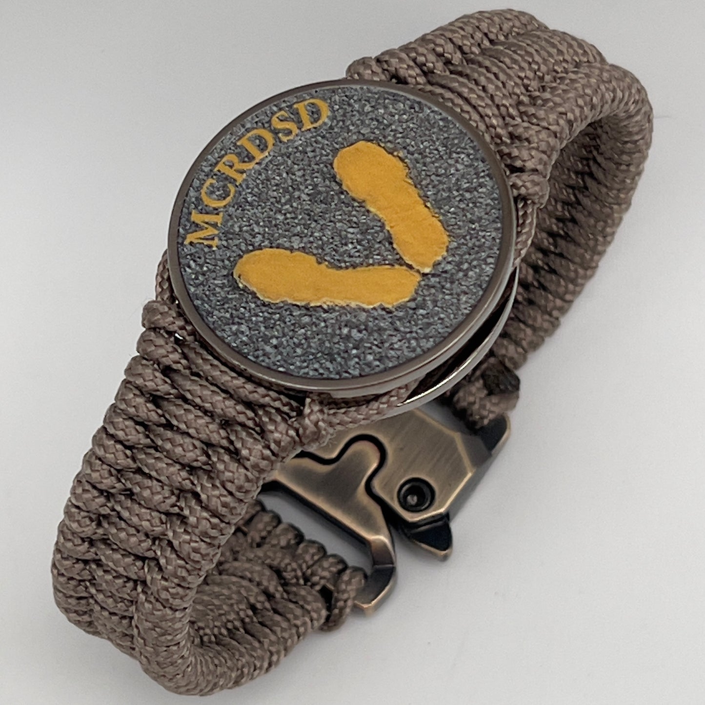Thin, narrow, flexible and comfortable braided United States Marine Corps Recruit Depot Yellow Footprints bracelet featuring a black nickel plated UV enamel painted medallion replica of an actual set of USMC recruit depot footprints, a gold Cobra style tactical buckle and woven with tan Type-1 paracord.
