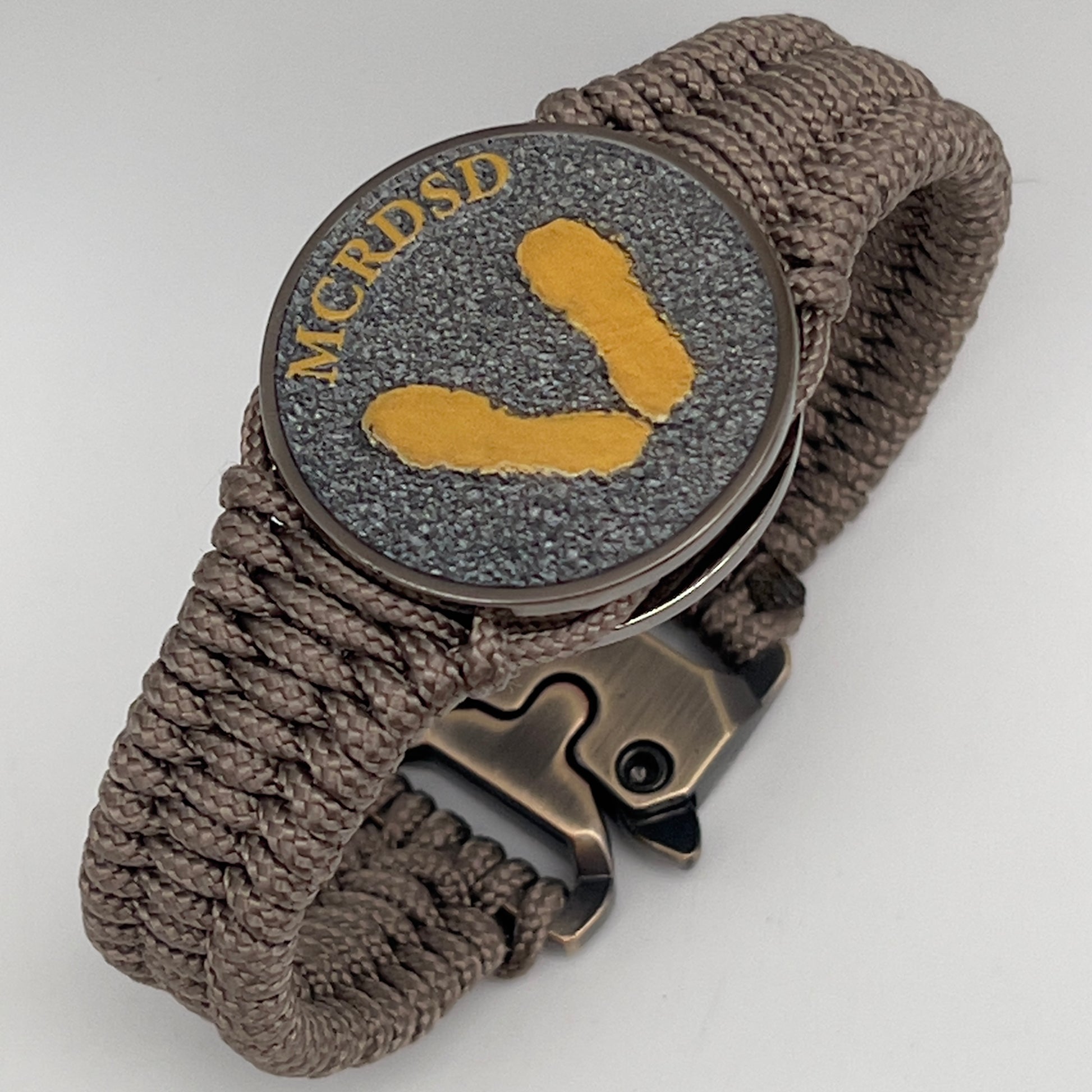 Thin, narrow, flexible and comfortable braided United States Marine Corps Recruit Depot Yellow Footprints bracelet featuring a black nickel plated UV enamel painted medallion replica of an actual set of USMC recruit depot footprints, a gold Cobra style tactical buckle and woven with tan Type-1 paracord.