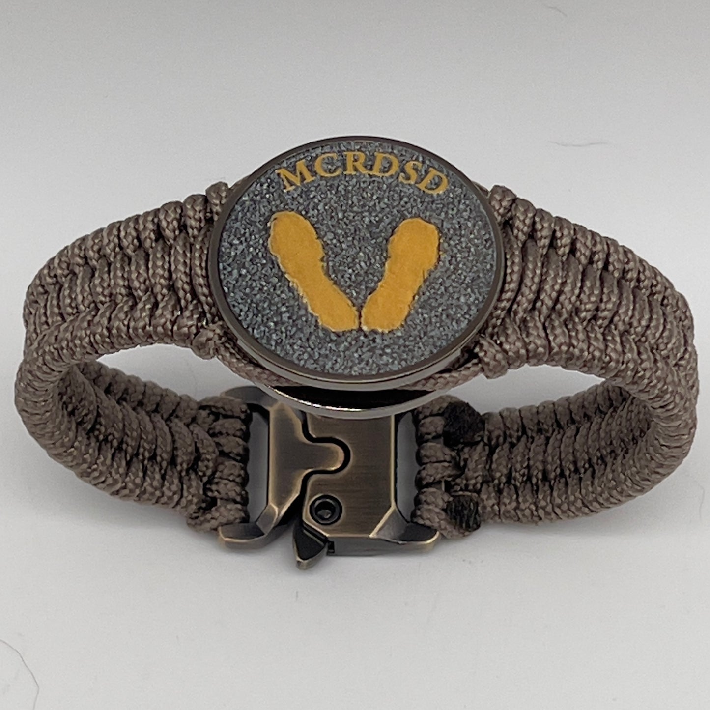 Thin, narrow, flexible and comfortable braided United States Marine Corps Recruit Depot Yellow Footprints bracelet featuring a black nickel plated UV enamel painted medallion replica of an actual set of USMC recruit depot footprints, a gold Cobra style tactical buckle and woven with tan Type-1 paracord.