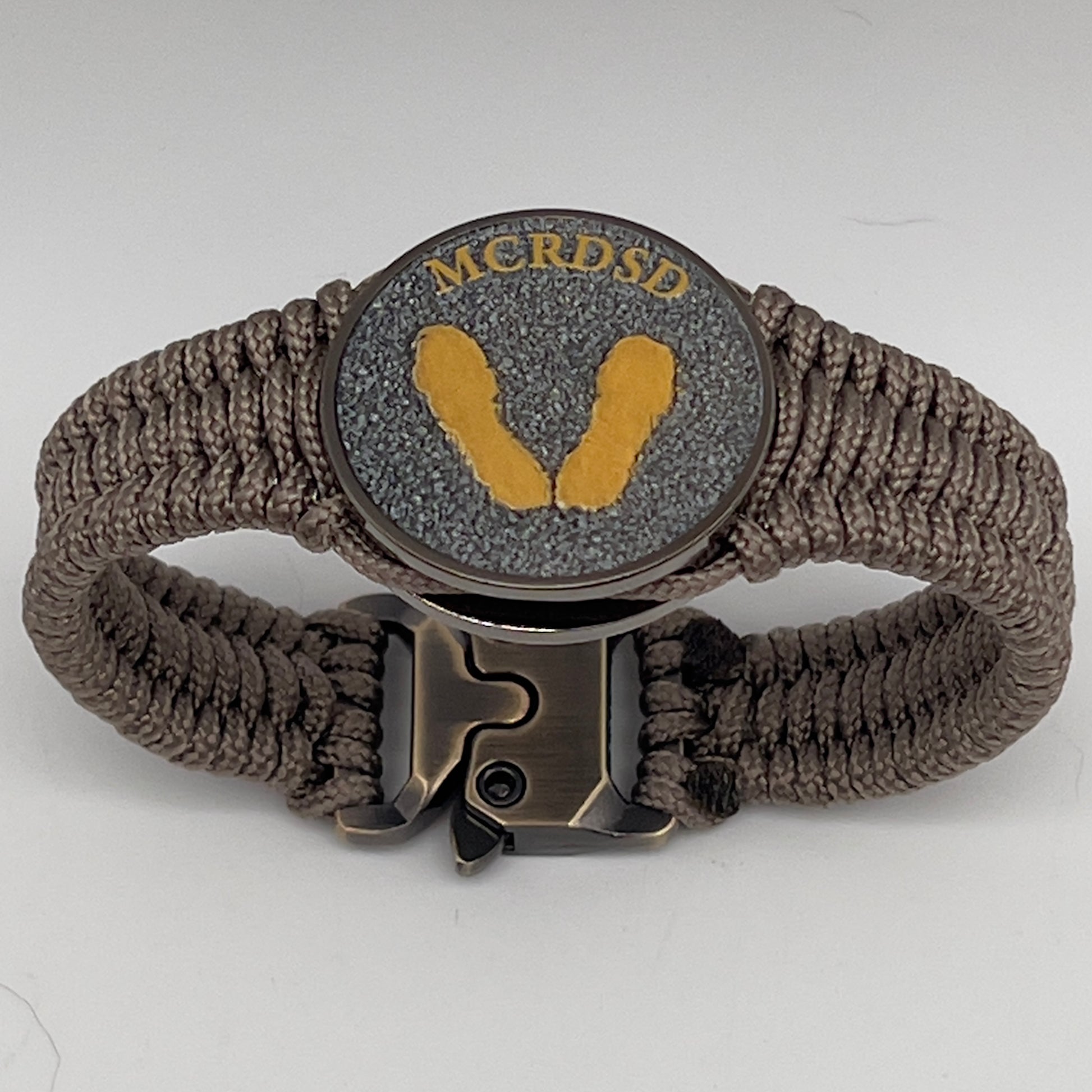 Thin, narrow, flexible and comfortable braided United States Marine Corps Recruit Depot Yellow Footprints bracelet featuring a black nickel plated UV enamel painted medallion replica of an actual set of USMC recruit depot footprints, a gold Cobra style tactical buckle and woven with tan Type-1 paracord.