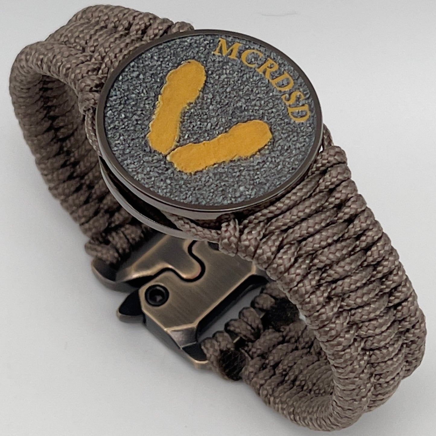 Thin, narrow, flexible and comfortable braided United States Marine Corps Recruit Depot Yellow Footprints bracelet featuring a black nickel plated UV enamel painted medallion replica of an actual set of USMC recruit depot footprints, a gold Cobra style tactical buckle and woven with tan Type-1 paracord.