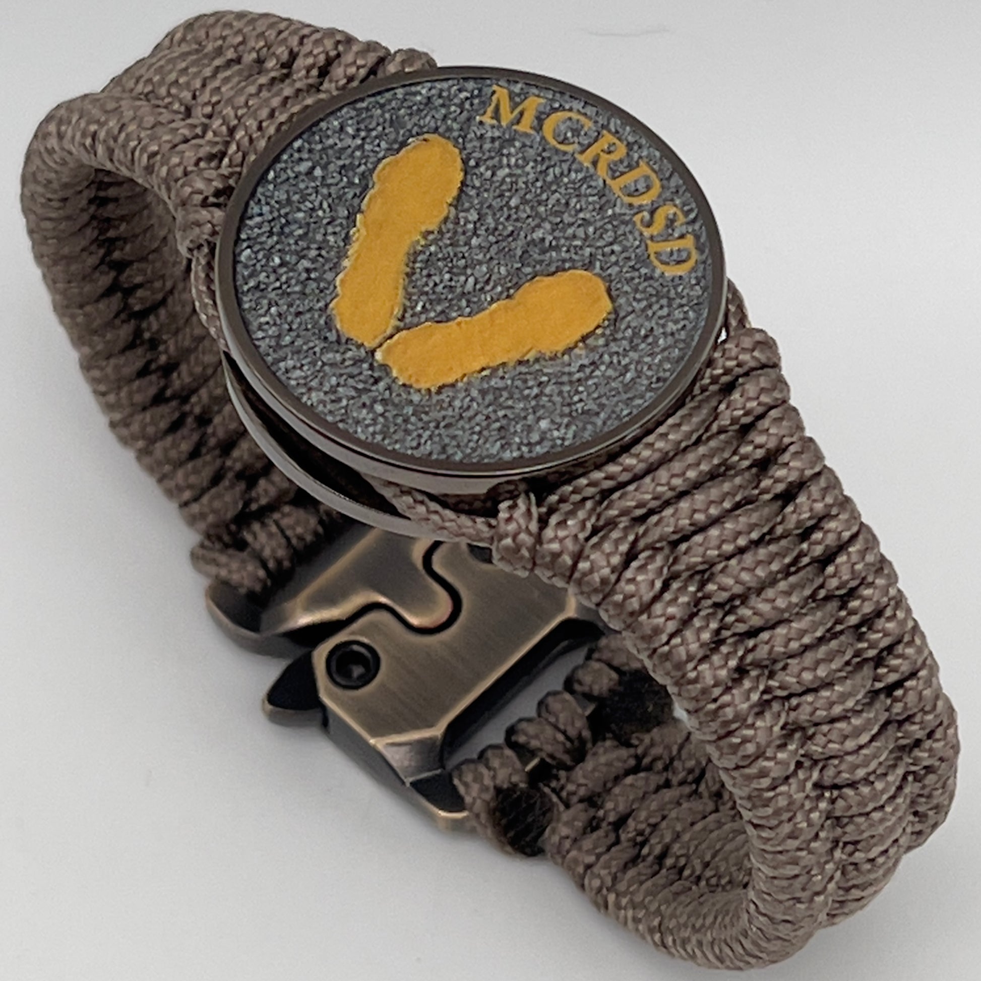 Thin, narrow, flexible and comfortable braided United States Marine Corps Recruit Depot Yellow Footprints bracelet featuring a black nickel plated UV enamel painted medallion replica of an actual set of USMC recruit depot footprints, a gold Cobra style tactical buckle and woven with tan Type-1 paracord.