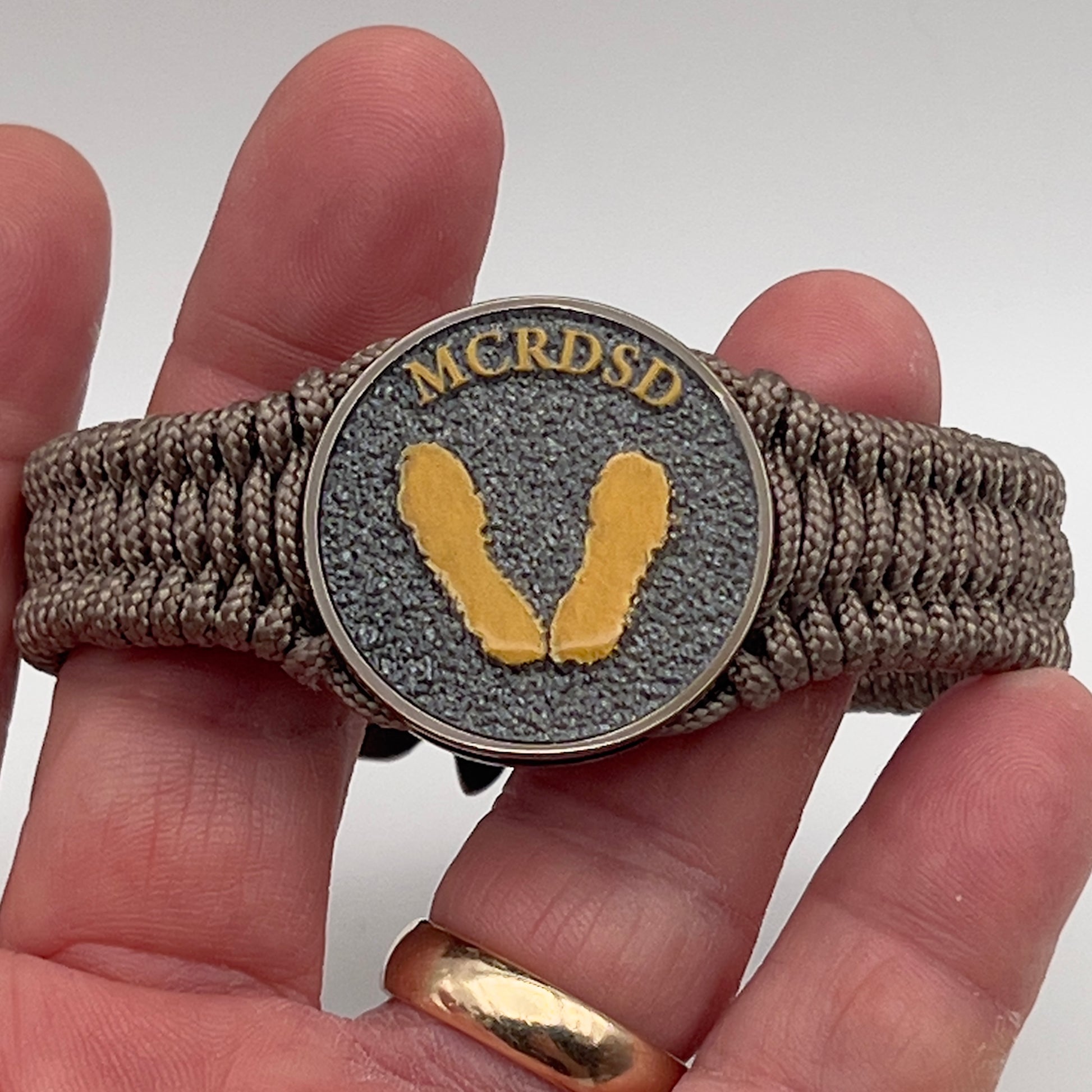 Thin, narrow, flexible and comfortable braided United States Marine Corps Recruit Depot Yellow Footprints bracelet featuring a black nickel plated UV enamel painted medallion replica of an actual set of USMC recruit depot footprints, a gold Cobra style tactical buckle and woven with tan Type-1 paracord.