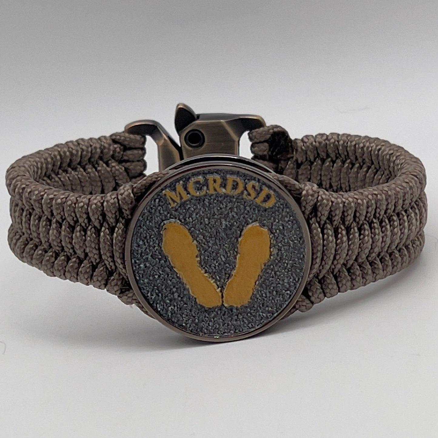 Thin, narrow, flexible and comfortable braided United States Marine Corps Recruit Depot Yellow Footprints bracelet featuring a black nickel plated UV enamel painted medallion replica of an actual set of USMC recruit depot footprints, a gold Cobra style tactical buckle and woven with tan Type-1 paracord.