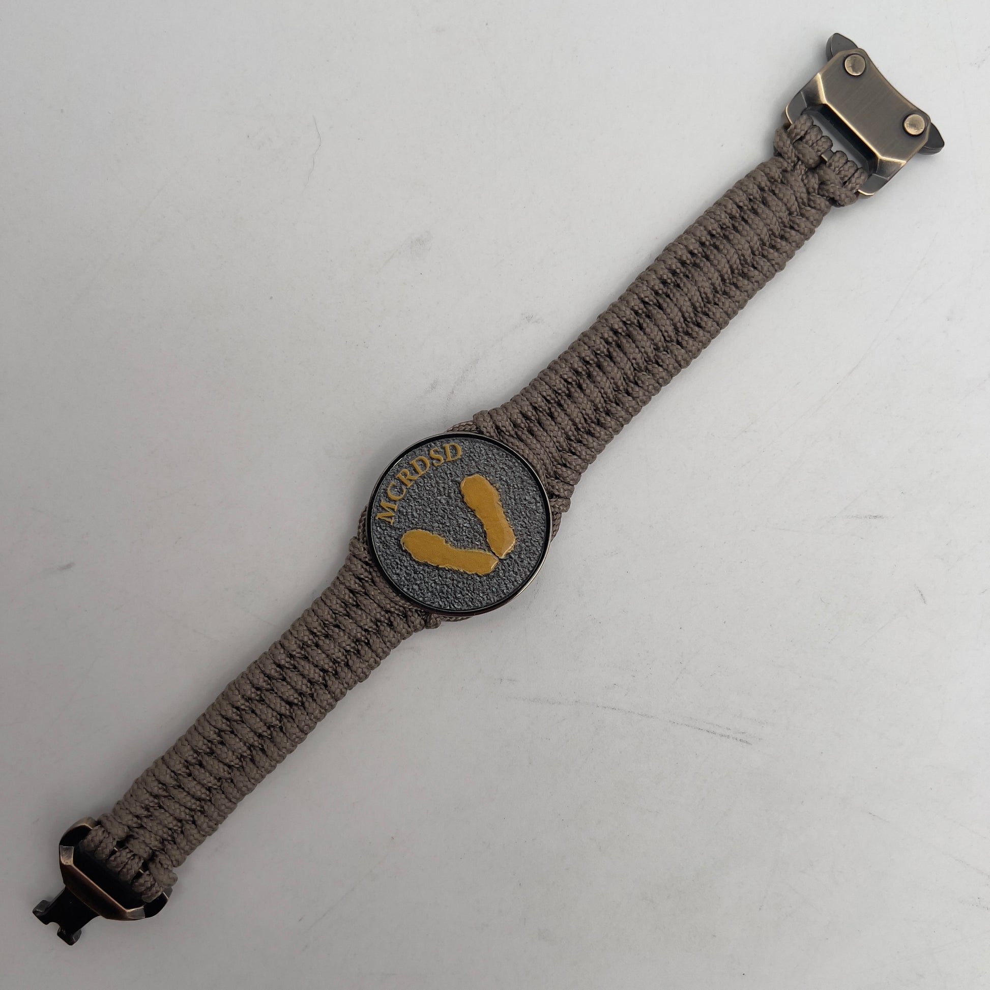 Thin, narrow, flexible and comfortable braided United States Marine Corps Recruit Depot Yellow Footprints bracelet featuring a black nickel plated UV enamel painted medallion replica of an actual set of USMC recruit depot footprints, a gold Cobra style tactical buckle and woven with tan Type-1 paracord.