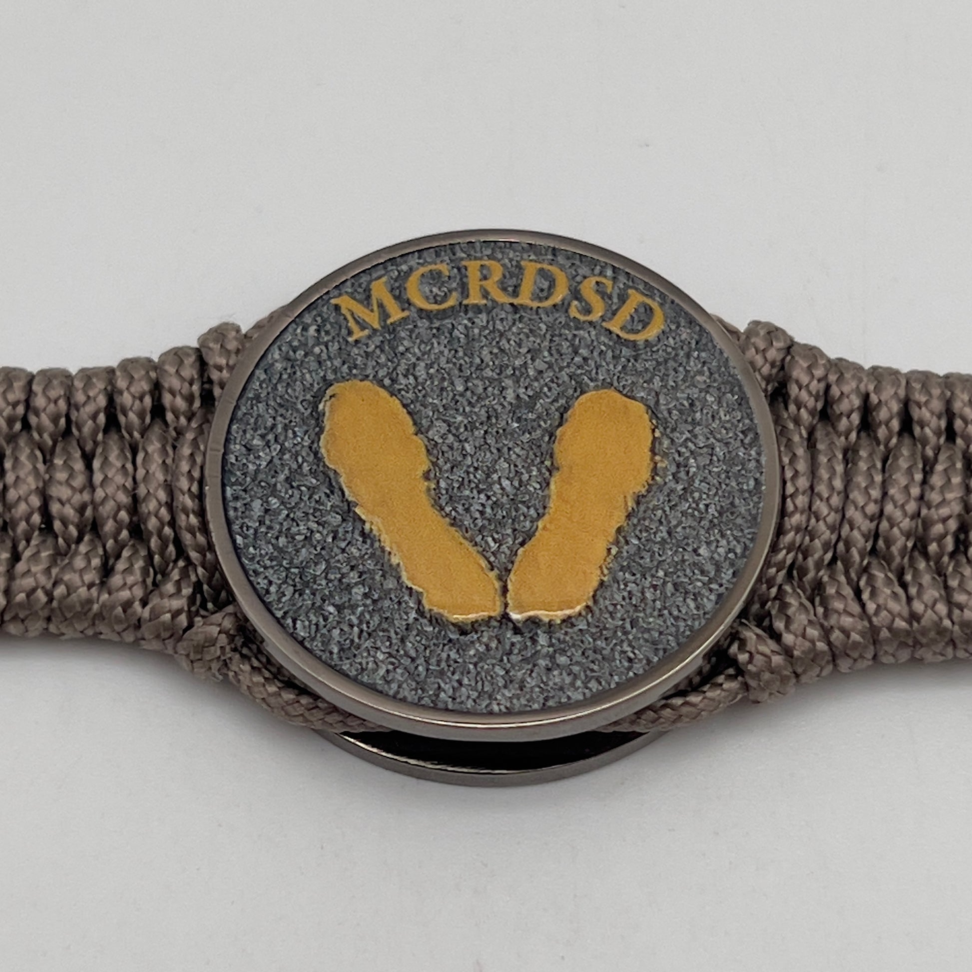 Thin, narrow, flexible and comfortable braided United States Marine Corps Recruit Depot Yellow Footprints bracelet featuring a black nickel plated UV enamel painted medallion replica of an actual set of USMC recruit depot footprints, a gold Cobra style tactical buckle and woven with tan Type-1 paracord.