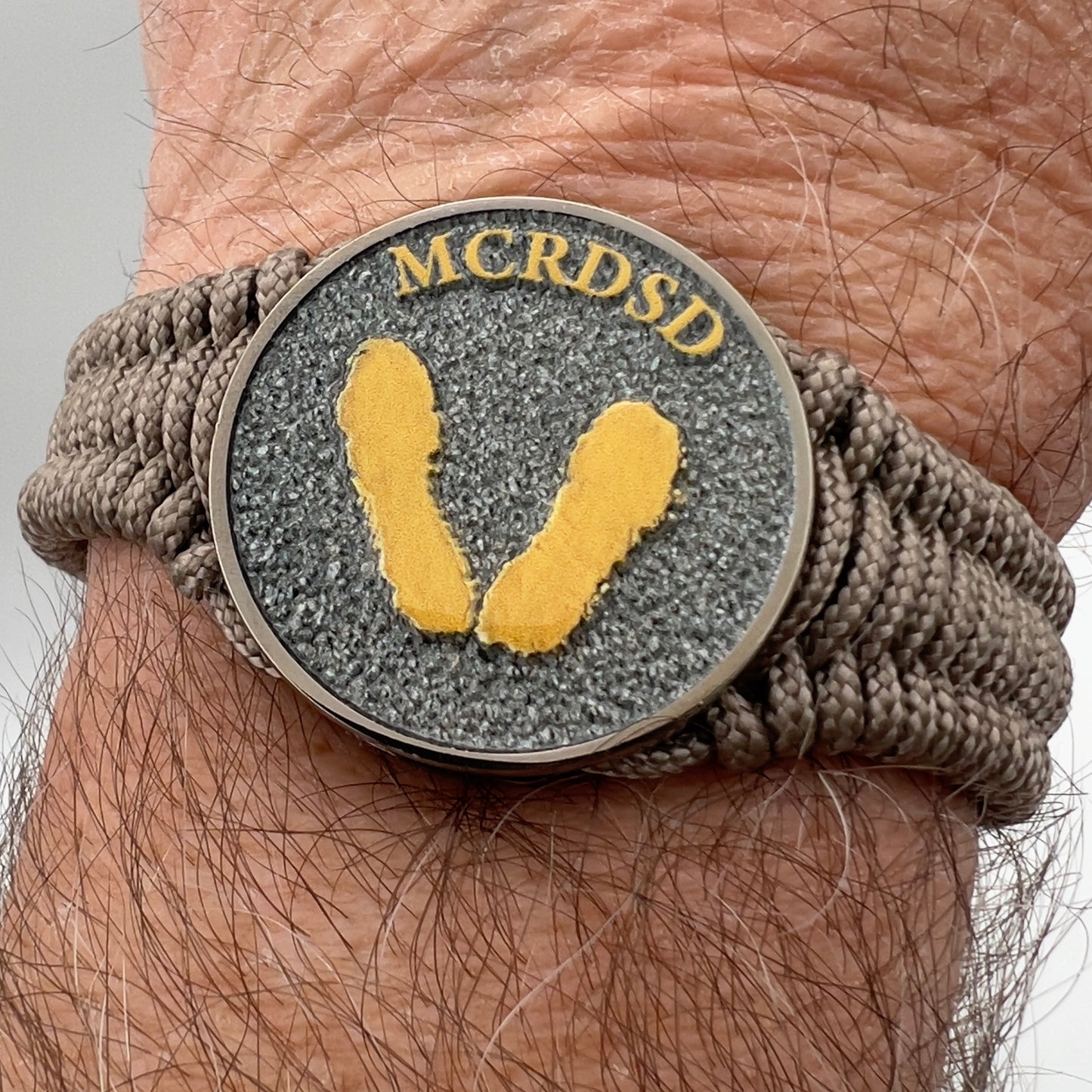 Thin, narrow, flexible and comfortable braided United States Marine Corps Recruit Depot Yellow Footprints bracelet featuring a black nickel plated UV enamel painted medallion replica of an actual set of USMC recruit depot footprints, a gold Cobra style tactical buckle and woven with tan Type-1 paracord.