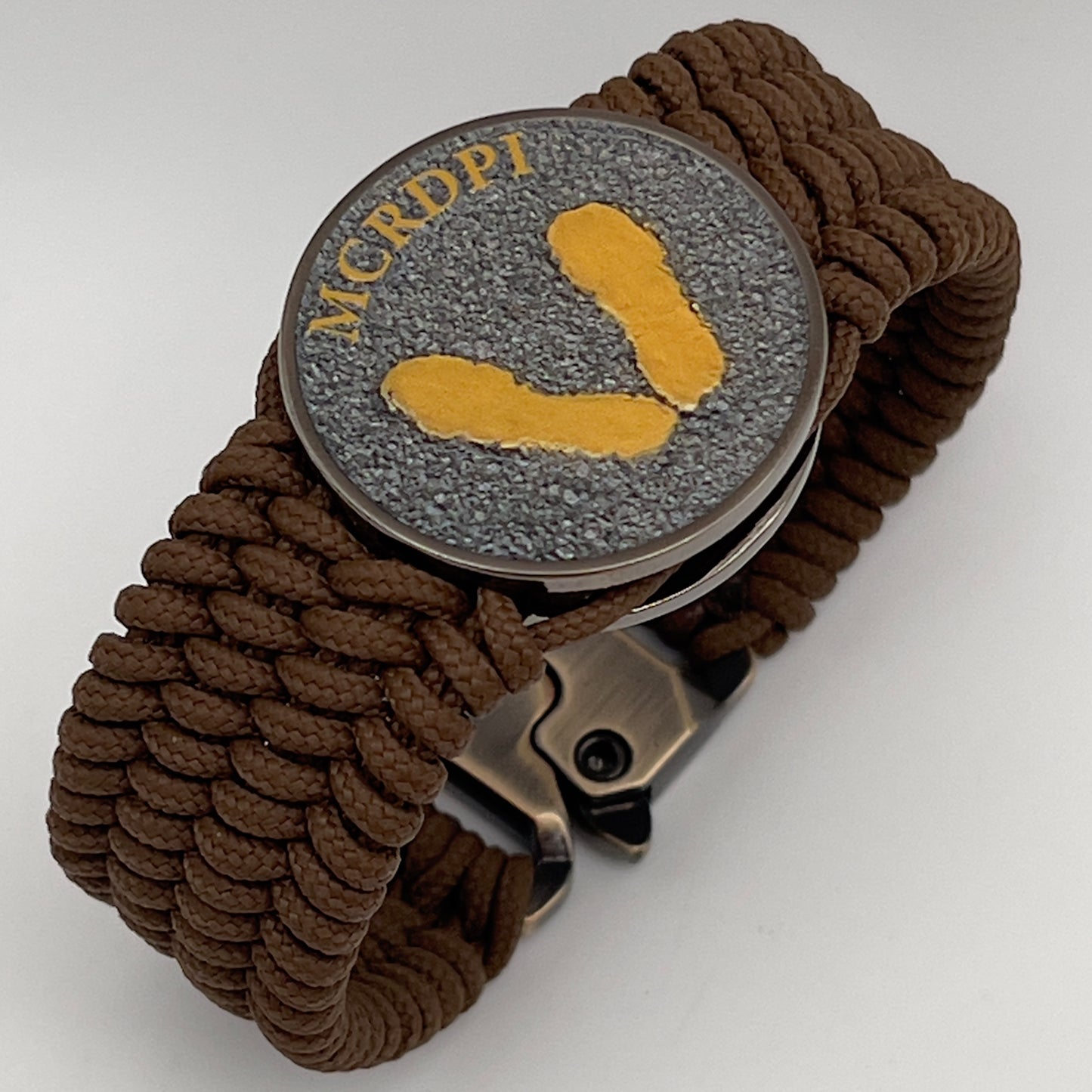 Thin, wide, flexible and comfortable braided United States Marine Corps Recruit Depot Yellow Footprints bracelet featuring a black nickel plated UV enamel painted medallion replica of an actual set of USMC footprints, a gold Cobra style tactical buckle and woven with Coyote Brown Type-1 paracord.