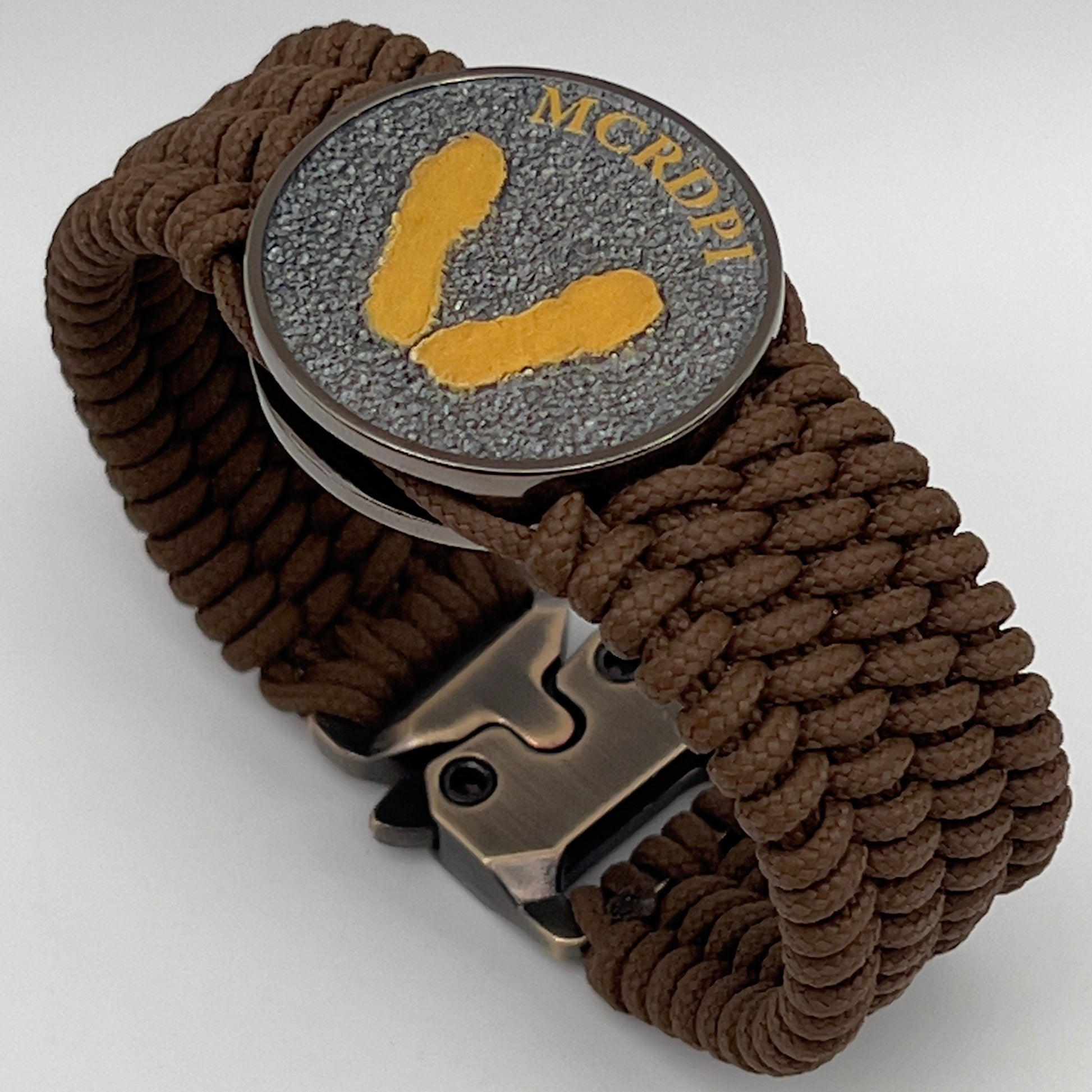 Thin, wide, flexible and comfortable braided United States Marine Corps Recruit Depot Yellow Footprints bracelet featuring a black nickel plated UV enamel painted medallion replica of an actual set of USMC footprints, a gold Cobra style tactical buckle and woven with Coyote Brown Type-1 paracord.