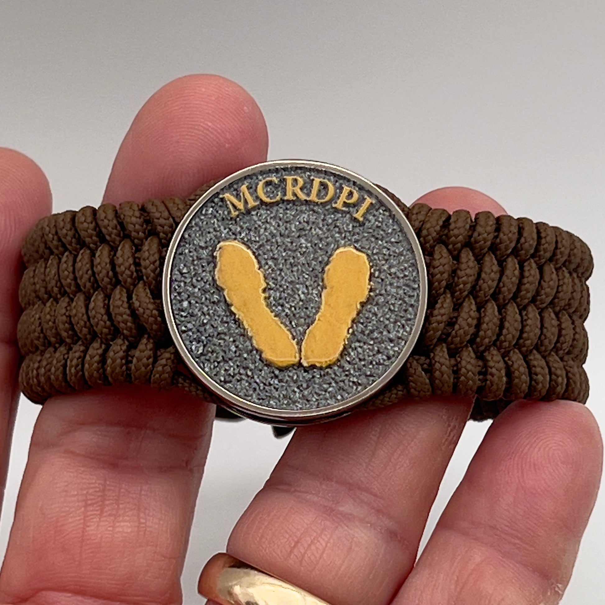 Thin, wide, flexible and comfortable braided United States Marine Corps Recruit Depot Yellow Footprints bracelet featuring a black nickel plated UV enamel painted medallion replica of an actual set of USMC footprints, a gold Cobra style tactical buckle and woven with Coyote Brown Type-1 paracord.