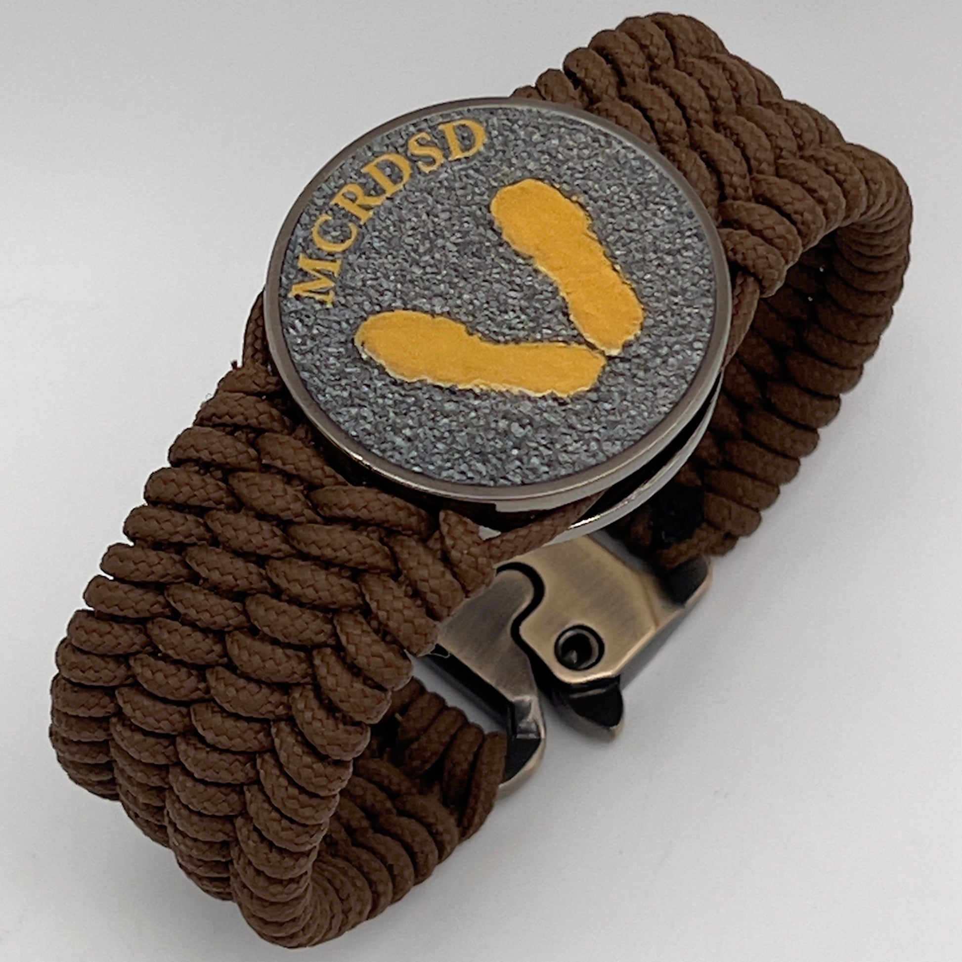 Thin, wide, flexible and comfortable braided United States Marine Corps Recruit Depot Yellow Footprints bracelet featuring a black nickel plated UV enamel painted medallion replica of an actual set of USMC footprints, a gold Cobra style tactical buckle and woven with Coyote Brown Type-1 paracord.