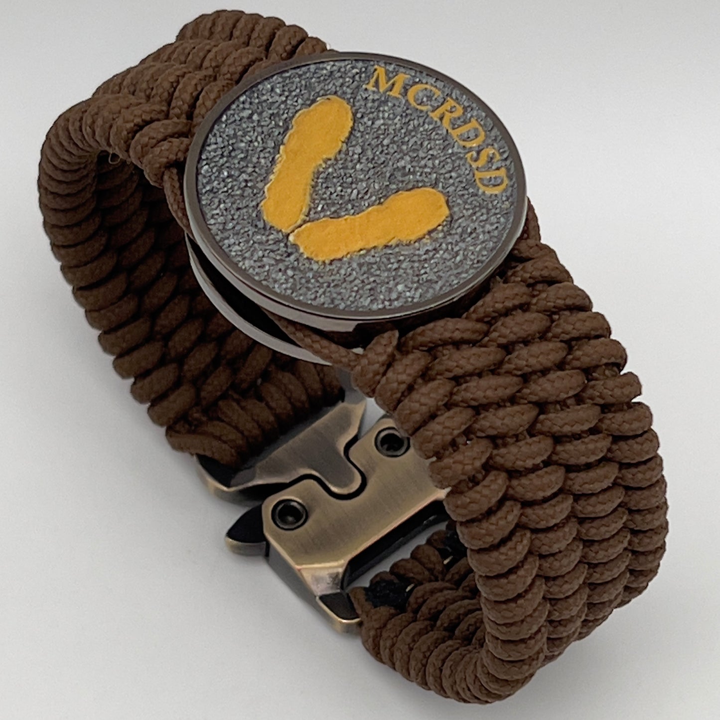 Thin, wide, flexible and comfortable braided United States Marine Corps Recruit Depot Yellow Footprints bracelet featuring a black nickel plated UV enamel painted medallion replica of an actual set of USMC footprints, a gold Cobra style tactical buckle and woven with Coyote Brown Type-1 paracord.