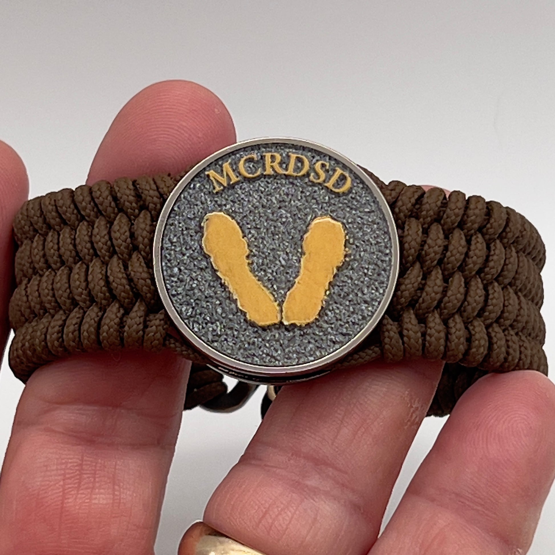 Thin, wide, flexible and comfortable braided United States Marine Corps Recruit Depot Yellow Footprints bracelet featuring a black nickel plated UV enamel painted medallion replica of an actual set of USMC footprints, a gold Cobra style tactical buckle and woven with Coyote Brown Type-1 paracord.