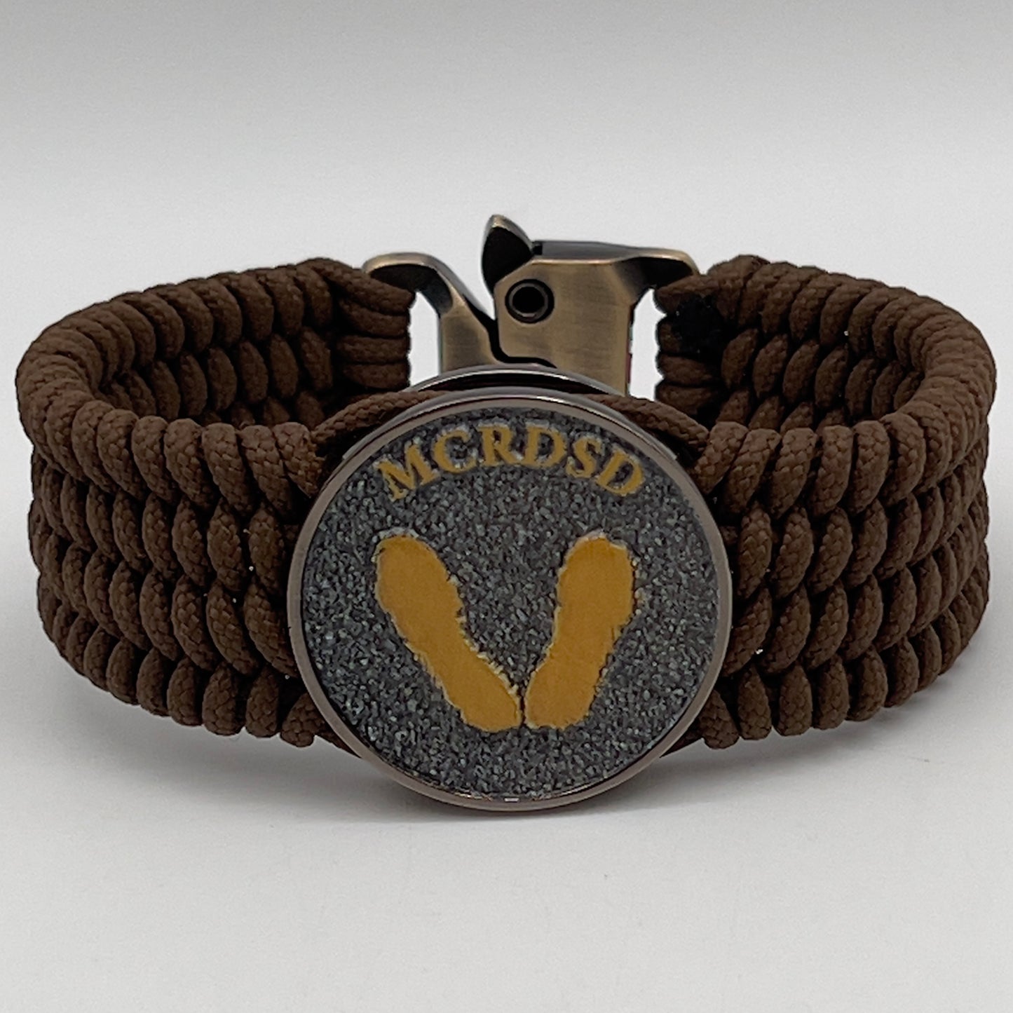 Thin, wide, flexible and comfortable braided United States Marine Corps Recruit Depot Yellow Footprints bracelet featuring a black nickel plated UV enamel painted medallion replica of an actual set of USMC footprints, a gold Cobra style tactical buckle and woven with Coyote Brown Type-1 paracord.
