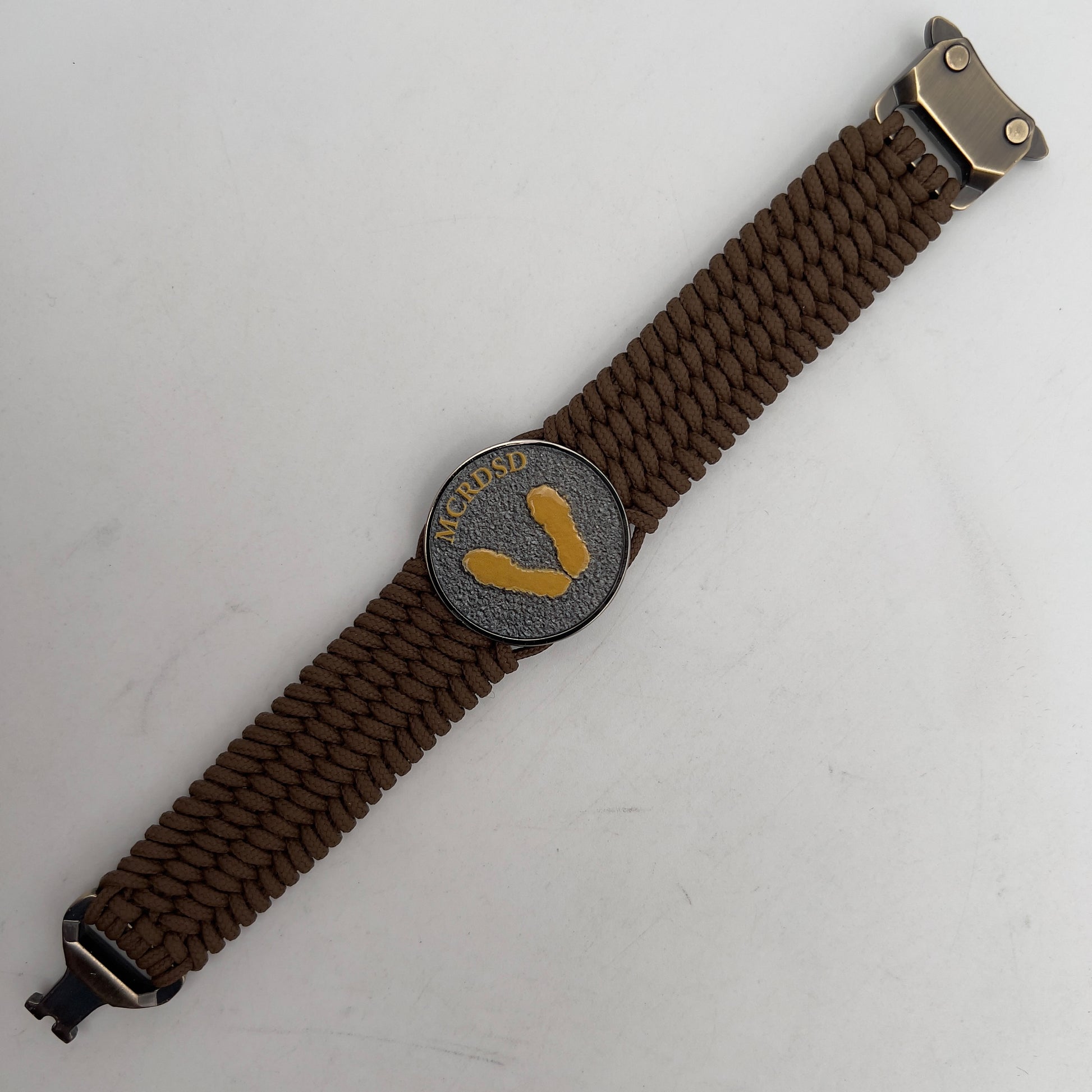 Thin, wide, flexible and comfortable braided United States Marine Corps Recruit Depot Yellow Footprints bracelet featuring a black nickel plated UV enamel painted medallion replica of an actual set of USMC footprints, a gold Cobra style tactical buckle and woven with Coyote Brown Type-1 paracord.
