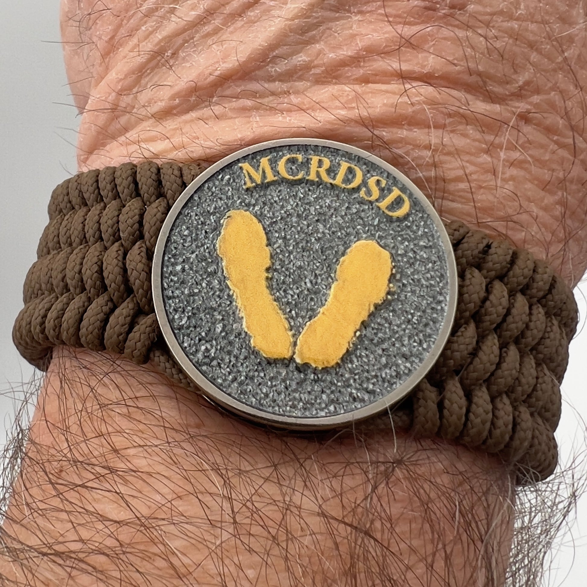 Thin, wide, flexible and comfortable braided United States Marine Corps Recruit Depot Yellow Footprints bracelet featuring a black nickel plated UV enamel painted medallion replica of an actual set of USMC footprints, a gold Cobra style tactical buckle and woven with Coyote Brown Type-1 paracord.