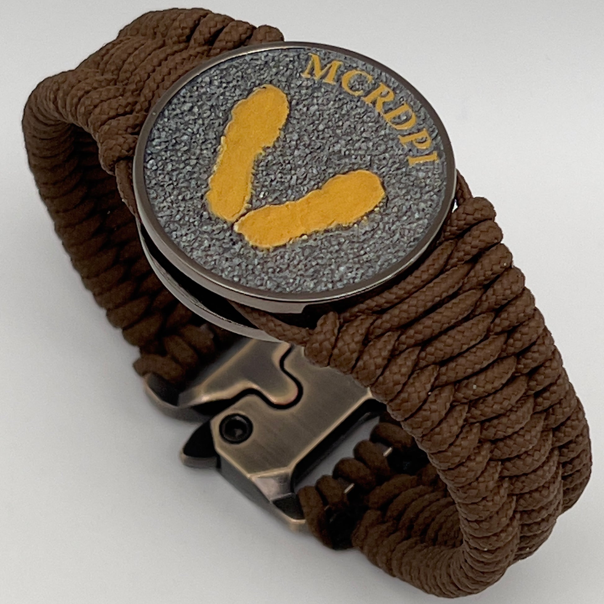 Thin, narrow, flexible and comfortable braided United States Marine Corps Recruit Depot Yellow Footprints bracelet featuring a black nickel plated UV enamel painted medallion replica of an actual set of USMC recruit depot footprints, a gold Cobra style tactical buckle and woven with Coyote Brown type-1 paracord.