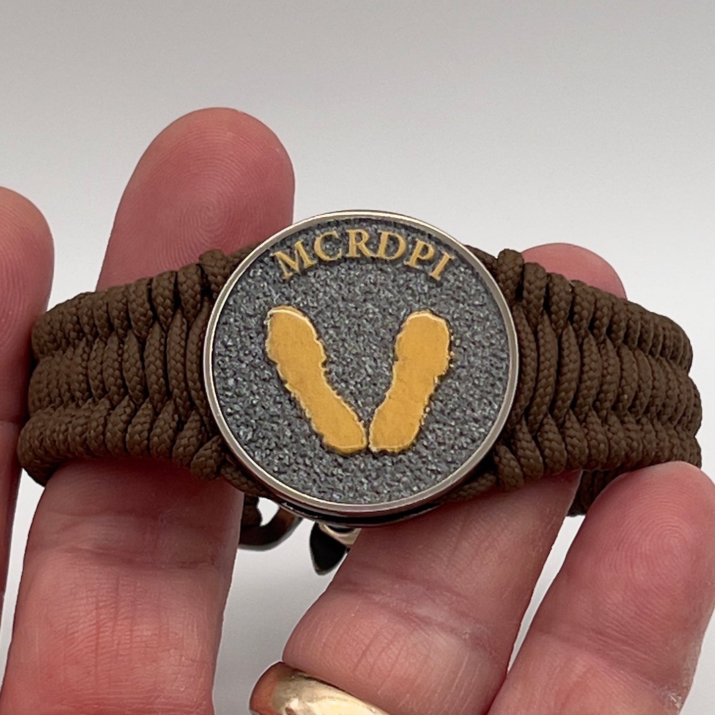 Thin, narrow, flexible and comfortable braided United States Marine Corps Recruit Depot Yellow Footprints bracelet featuring a black nickel plated UV enamel painted medallion replica of an actual set of USMC recruit depot footprints, a gold Cobra style tactical buckle and woven with Coyote Brown type-1 paracord.
