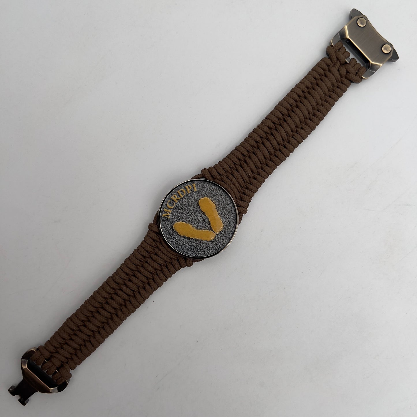 Thin, narrow, flexible and comfortable braided United States Marine Corps Recruit Depot Yellow Footprints bracelet featuring a black nickel plated UV enamel painted medallion replica of an actual set of USMC recruit depot footprints, a gold Cobra style tactical buckle and woven with Coyote Brown type-1 paracord.