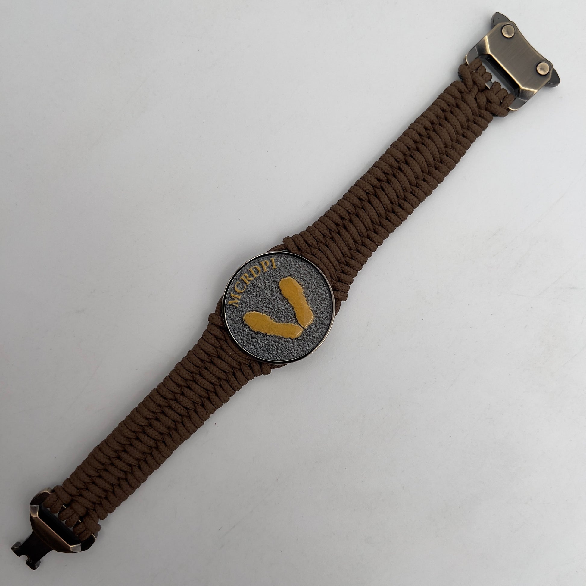 Thin, narrow, flexible and comfortable braided United States Marine Corps Recruit Depot Yellow Footprints bracelet featuring a black nickel plated UV enamel painted medallion replica of an actual set of USMC recruit depot footprints, a gold Cobra style tactical buckle and woven with Coyote Brown type-1 paracord.