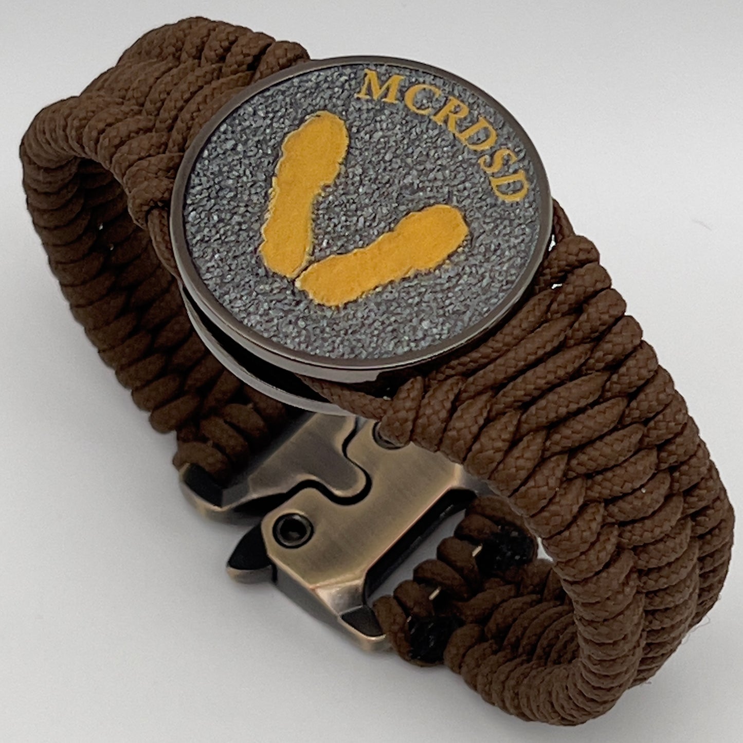 Thin, narrow, flexible and comfortable braided United States Marine Corps Recruit Depot Yellow Footprints bracelet featuring a black nickel plated UV enamel painted medallion replica of an actual set of USMC recruit depot footprints, a gold Cobra style tactical buckle and woven with Coyote Brown type-1 paracord.